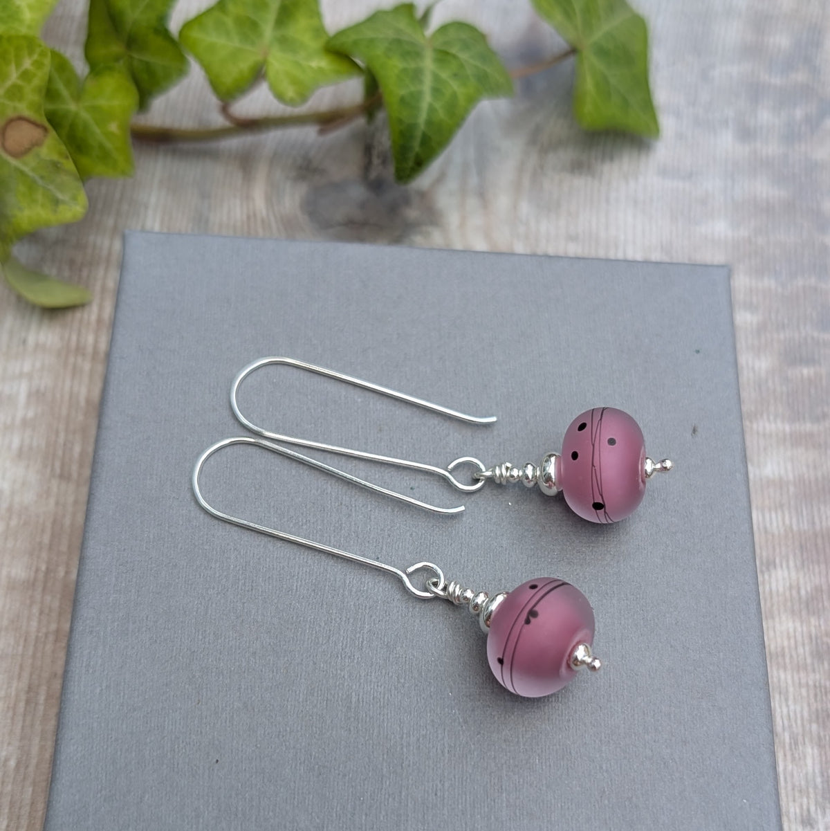 pink scribbles lampwork glass long silver earrings handmade from recycled Sterling Silver with frosted pink glass beads, displayed on a grey background.
