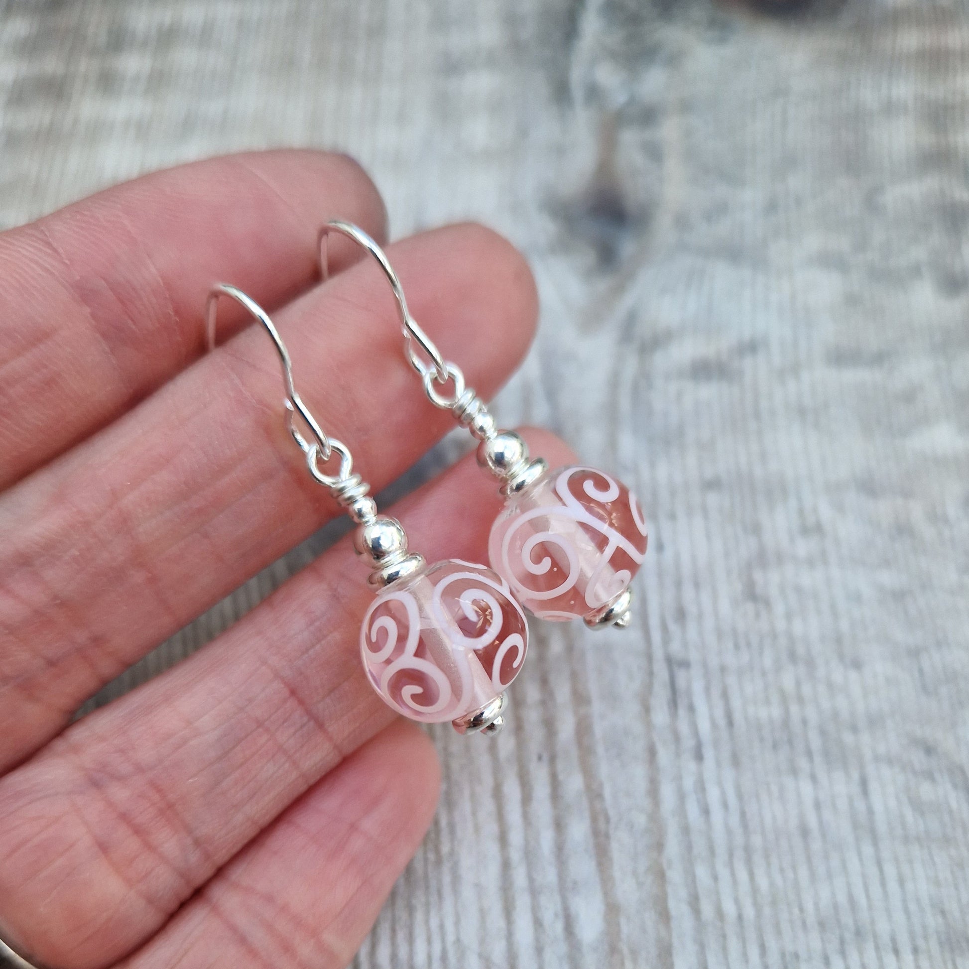 Each earring is a pink translucent lampwork bead suspended from silver earring wire via small, silver concentric spheres. Each bead has been inlaid with pale pink scroll patterns.