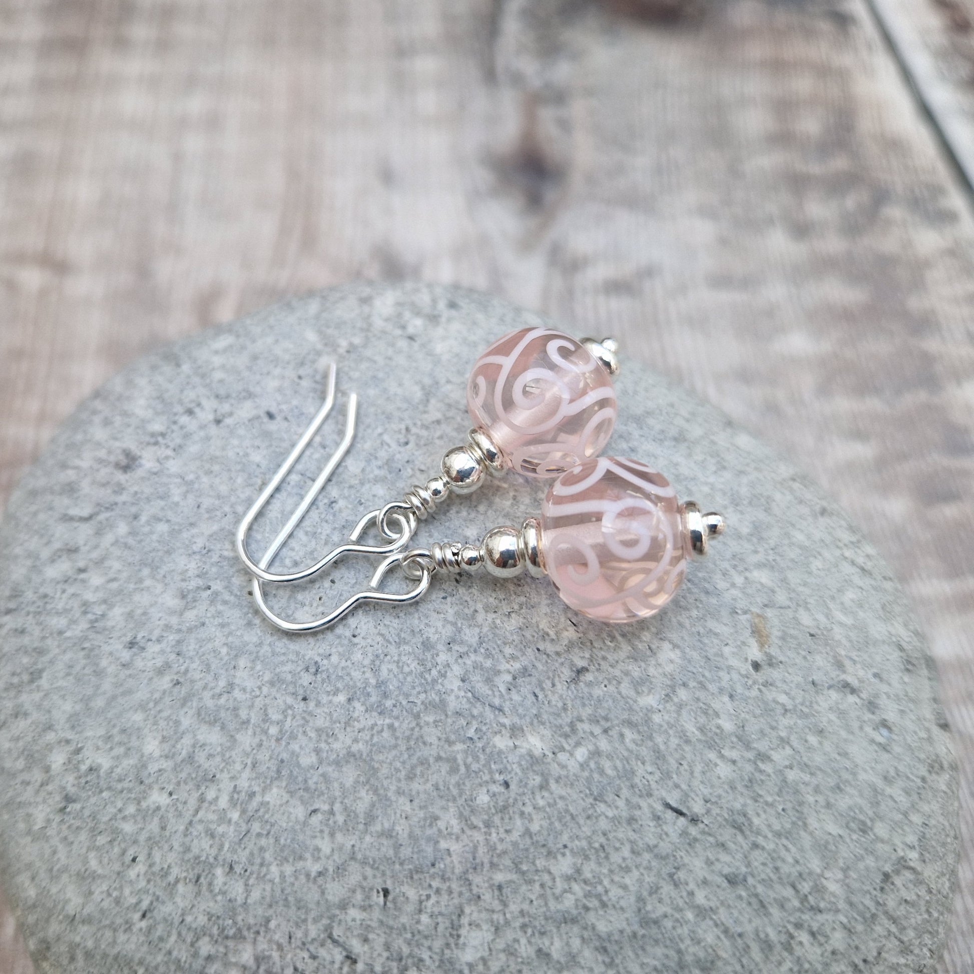 Each earring is a pink translucent lampwork bead suspended from silver earring wire via small, silver concentric spheres. Each bead has been inlaid with pale pink scroll patterns.