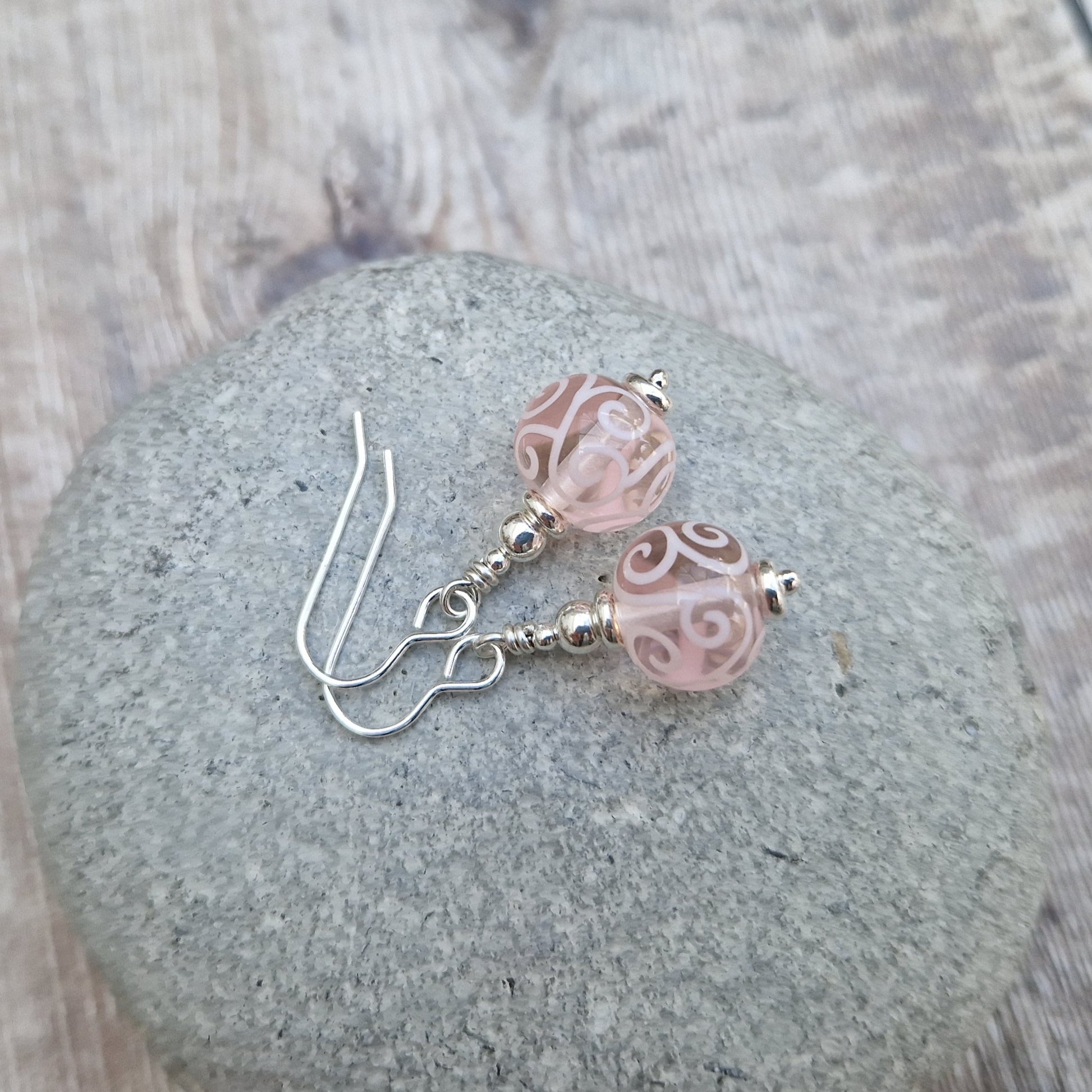 Each earring is a pink translucent lampwork bead suspended from silver earring wire via small, silver concentric spheres. Each bead has been inlaid with pale pink scroll patterns.