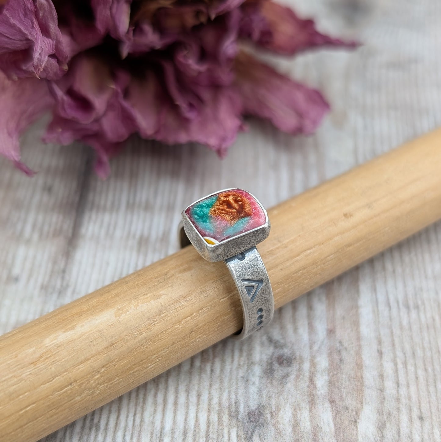 Rectangle shaped surfite stone with gloss finish mounted into oxidised Sterling Silver bezel and set on an oxidised silver band approx. 5mm wide. Ring band has been stamped with a geometric triangle and dot repeating pattern. The surfite has uneven flecks of pink, burnt orange and turquoise.