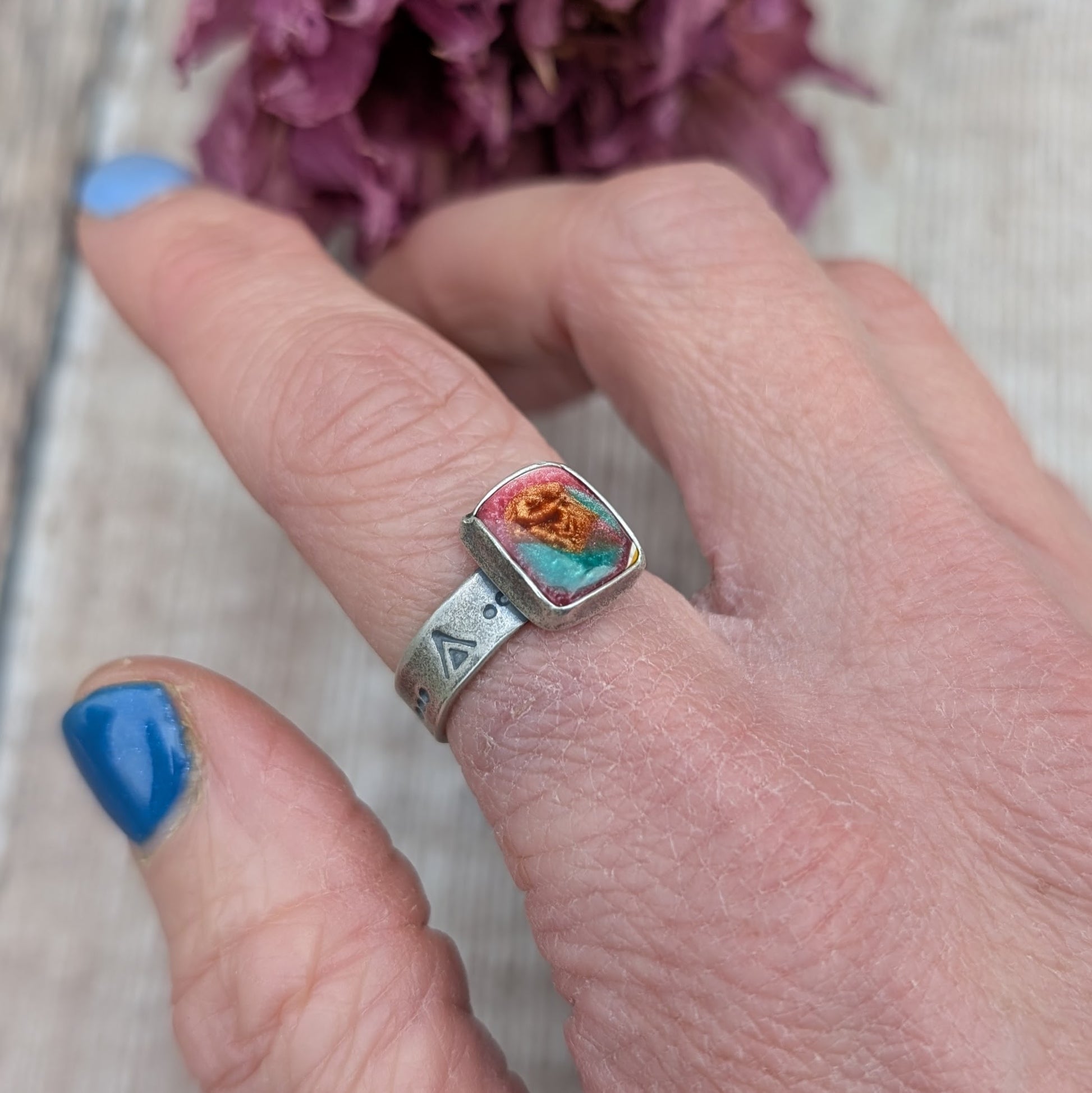 Rectangle shaped surfite stone with gloss finish mounted into oxidised Sterling Silver bezel and set on an oxidised silver band approx. 5mm wide. Ring band has been stamped with a geometric triangle and dot repeating pattern. The surfite has uneven flecks of pink, burnt orange and turquoise.