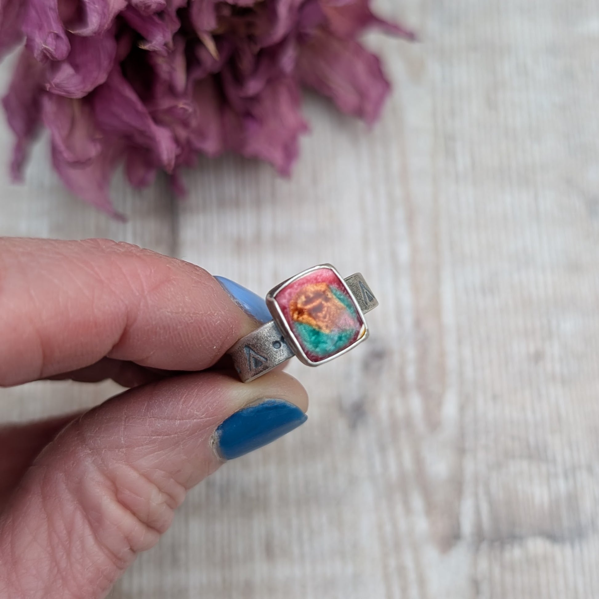 Rectangle shaped surfite stone with gloss finish mounted into oxidised Sterling Silver bezel and set on an oxidised silver band approx. 5mm wide. Ring band has been stamped with a geometric triangle and dot repeating pattern. The surfite has uneven flecks of pink, burnt orange and turquoise.