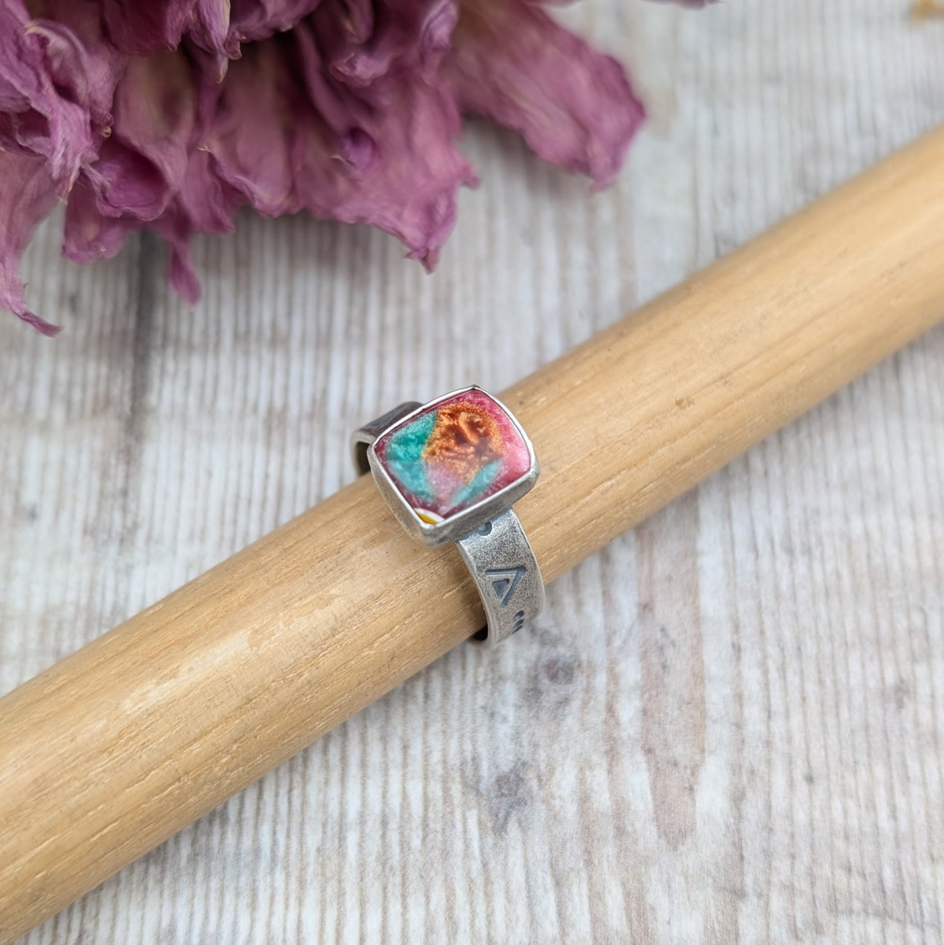 Rectangle shaped surfite stone with gloss finish mounted into oxidised Sterling Silver bezel and set on an oxidised silver band approx. 5mm wide. Ring band has been stamped with a geometric triangle and dot repeating pattern. The surfite has uneven flecks of pink, burnt orange and turquoise.