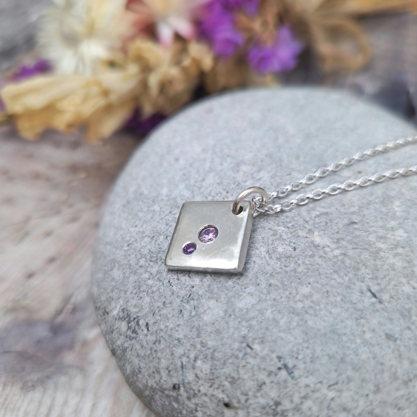 Sterling Silver off-set square pendant, smooth finish, slightly rounded long edges. Small round pink diamond-cut Cubic Zirconia stone deep set in the centre of the pendant with even smaller purple diamond-cut Cubic Zirconia stone set at bottom corner of the square. Small hole in top corner of off-set square, through which small silver circle links to silver chain.