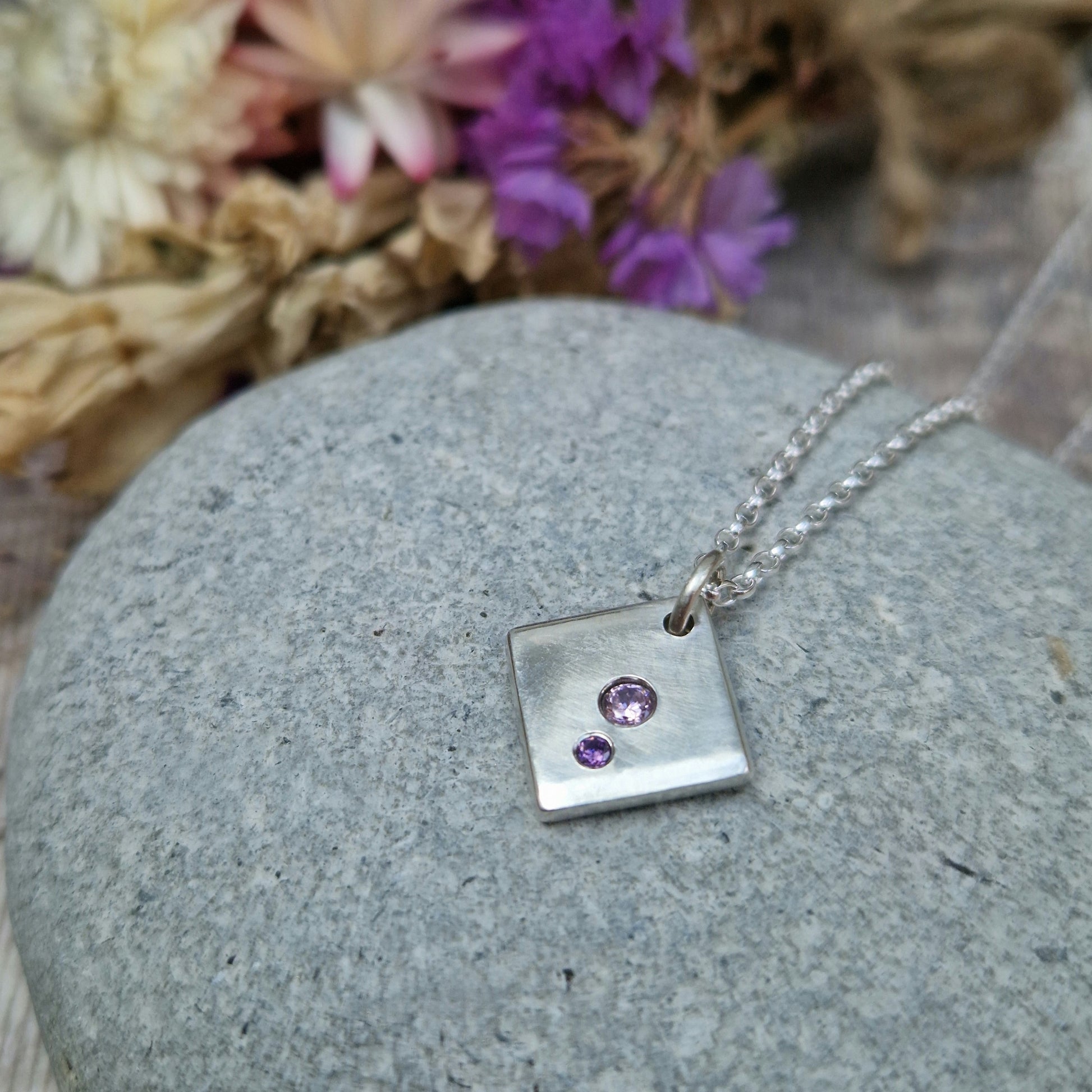 Sterling Silver off-set square pendant, smooth finish, slightly rounded long edges. Small round pink diamond-cut Cubic Zirconia stone deep set in the centre of the pendant with even smaller purple diamond-cut Cubic Zirconia stone set at bottom corner of the square. Small hole in top corner of off-set square, through which small silver circle links to silver chain.