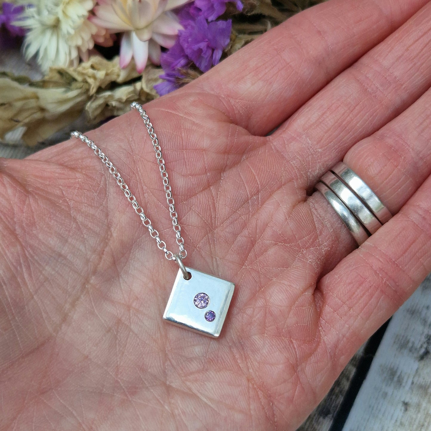 Sterling Silver off-set square pendant, smooth finish, slightly rounded long edges. Small round pink diamond-cut Cubic Zirconia stone deep set in the centre of the pendant with even smaller purple diamond-cut Cubic Zirconia stone set at bottom corner of the square. Small hole in top corner of off-set square, through which small silver circle links to silver chain.