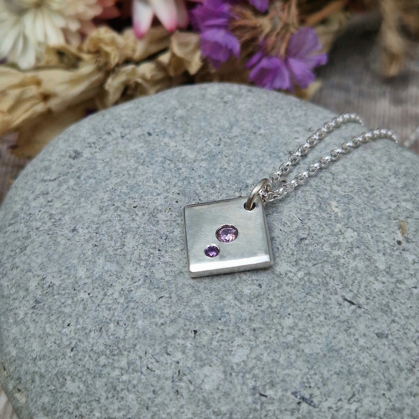 Sterling Silver off-set square pendant, smooth finish, slightly rounded long edges. Small round pink diamond-cut Cubic Zirconia stone deep set in the centre of the pendant with even smaller purple diamond-cut Cubic Zirconia stone set at bottom corner of the square. Small hole in top corner of off-set square, through which small silver circle links to silver chain.