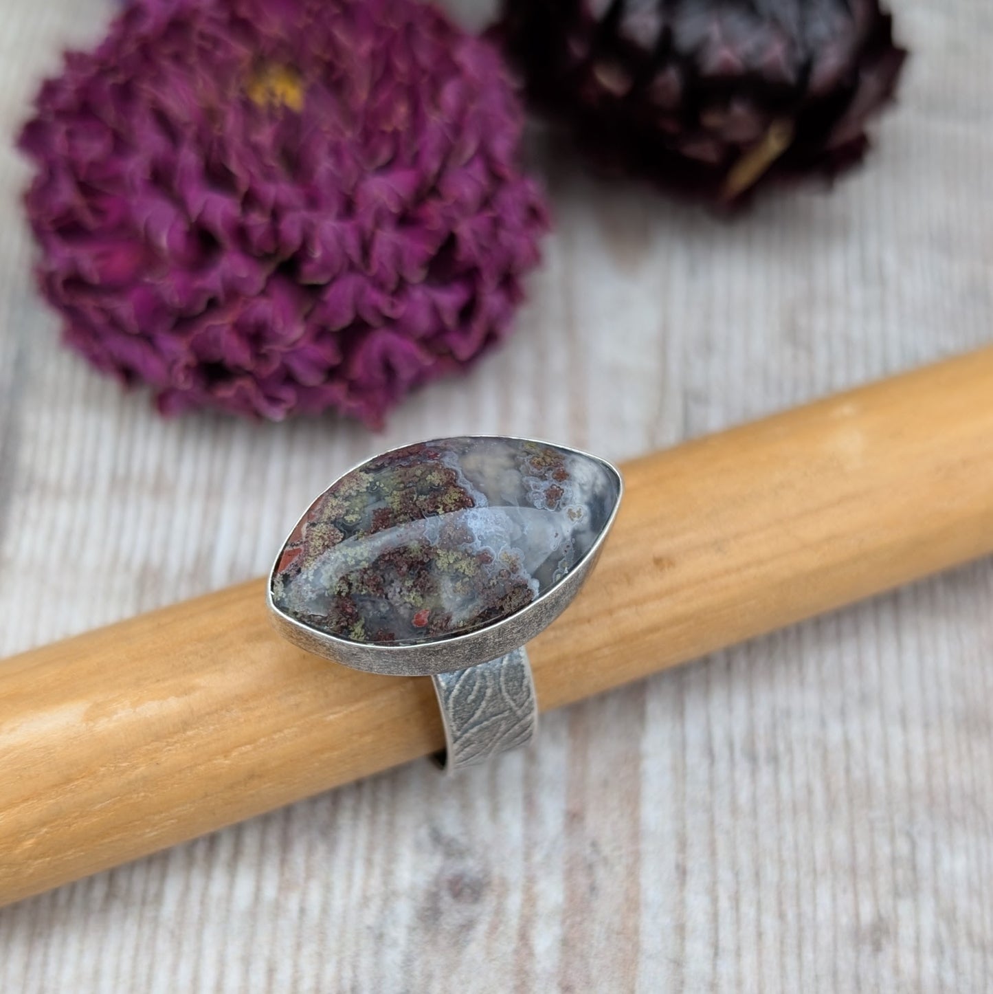 Silver Plume Agate Ring displayed on wooden holder with dried flowers in background, showing handmade silver band and plume agate stone