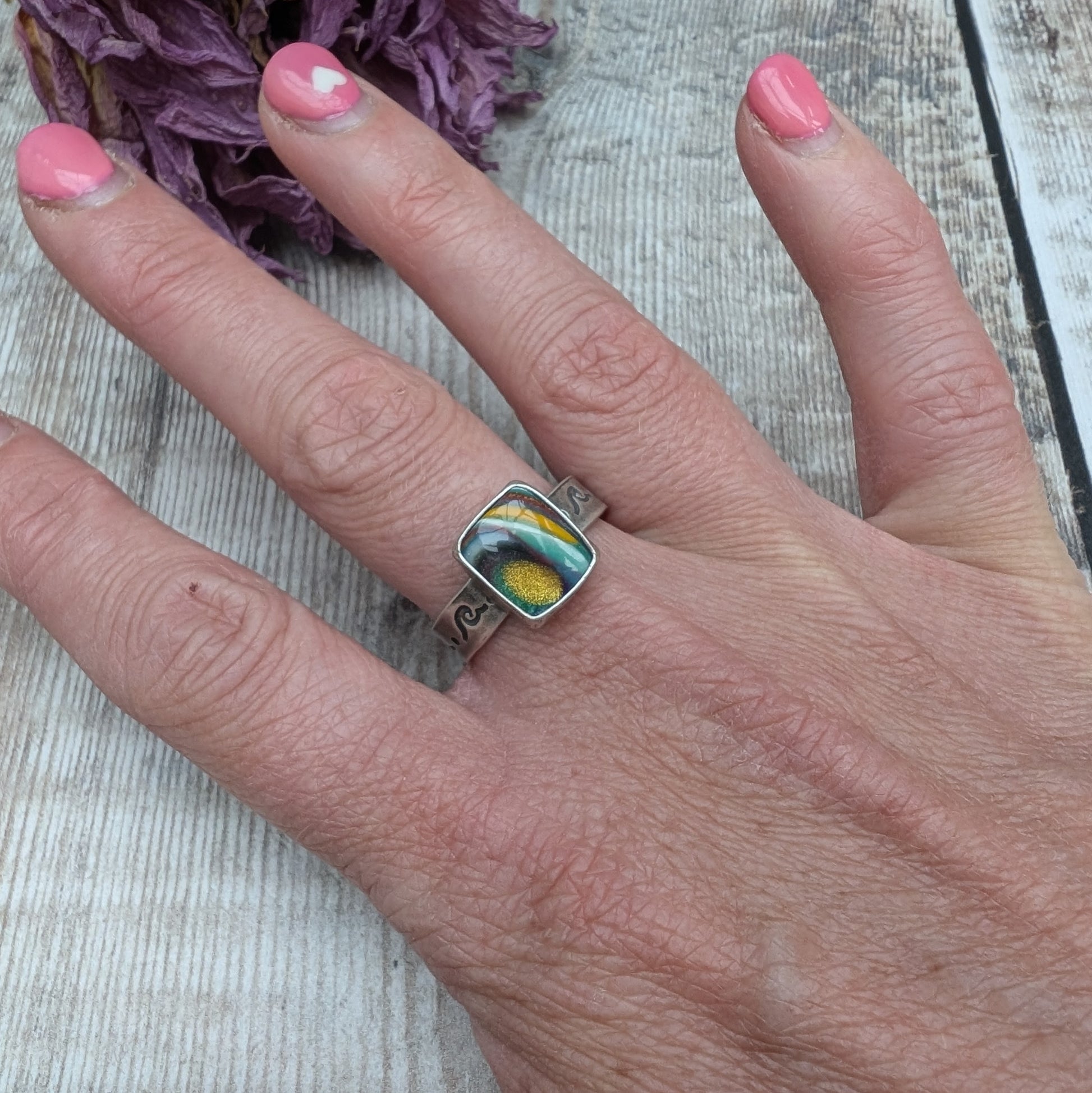 Rainbow Surfite stone measuring approx. 10mm x 12mm, mounted into oxidised Sterling Silver bezel and set on an oxidised silver band approx. 5mm wide. Ring band has been stamped with a wave and dot repeating pattern. The surfite has uneven swirls of red, yellow, aqua blue, black and a burst of sparkling gold in one corner.