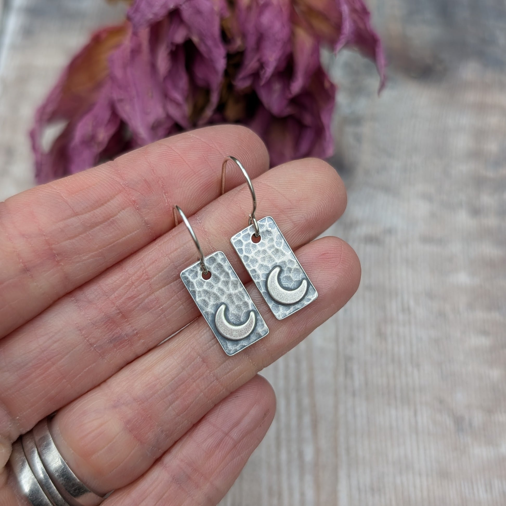 Hand holding a pair of oxidised rectangular earrings with a hammered texture and crescent moon details, with purple dried flowers in the background.