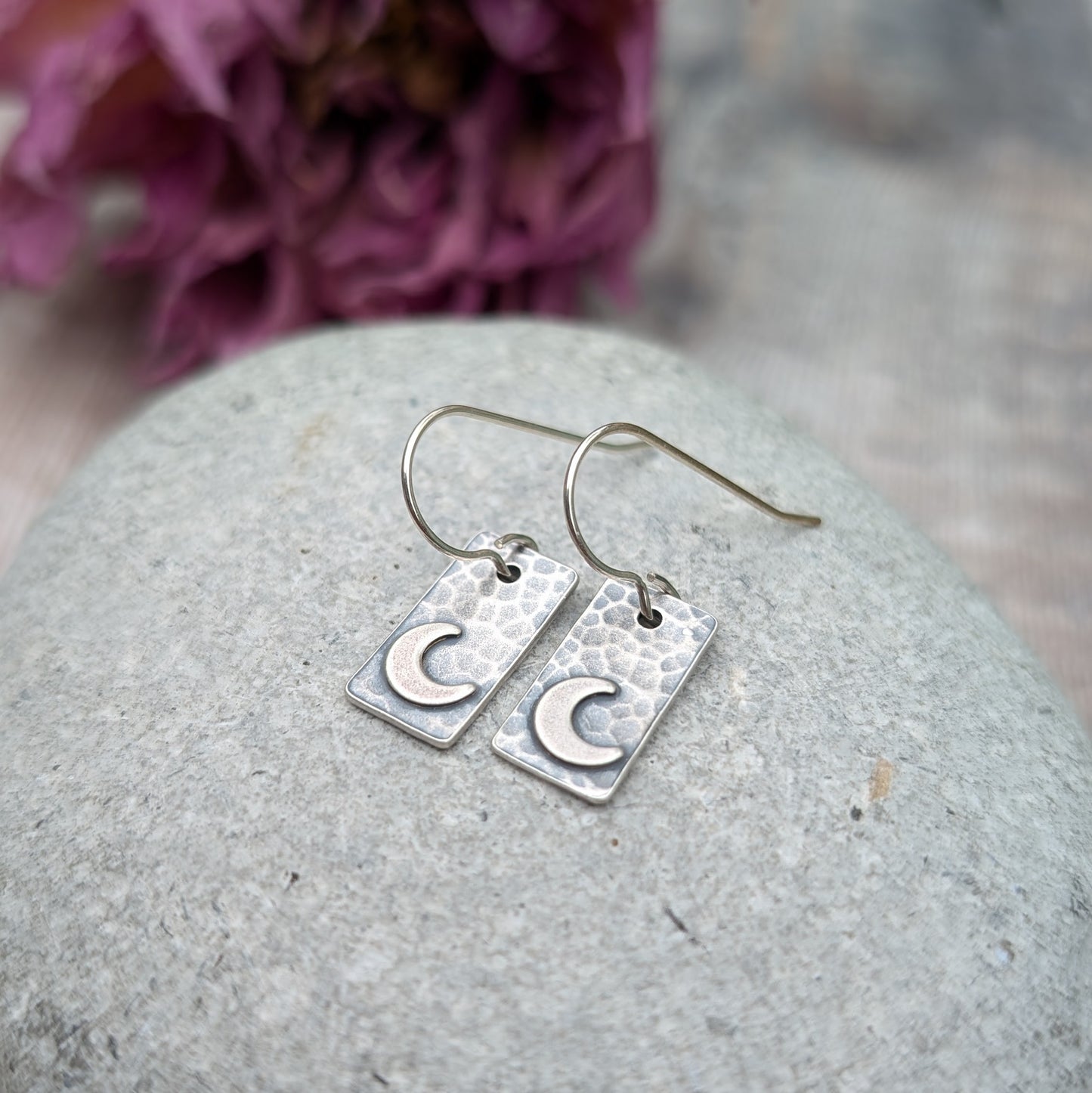 Pair of oxidised rectangular earrings with hammered texture and crescent moon details, resting on a grey stone background with purple dried flowers in the background.