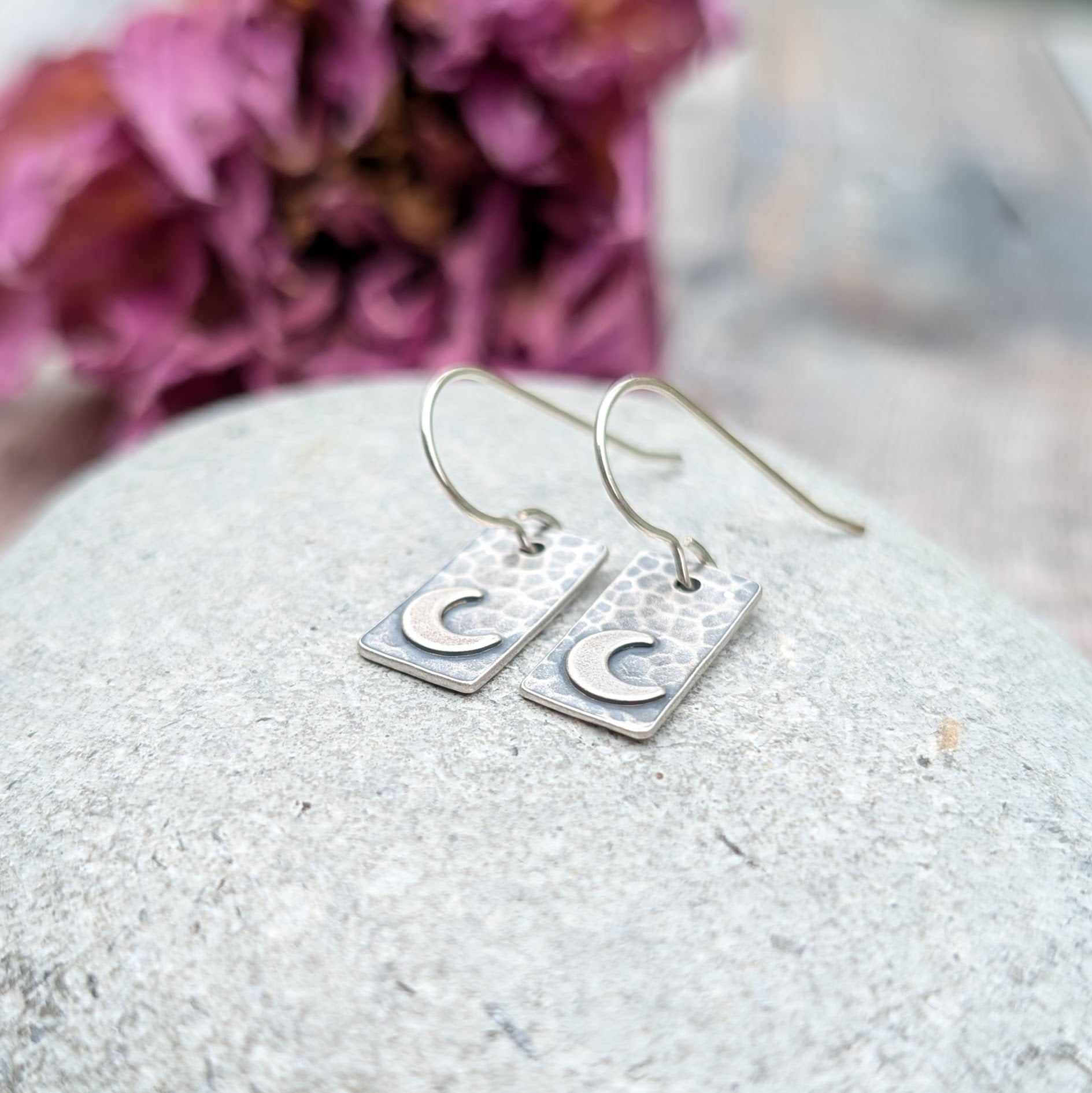 Pair of oxidised rectangular earrings with hammered texture and crescent moon details, resting on a grey stone background with purple dried flowers in the background.