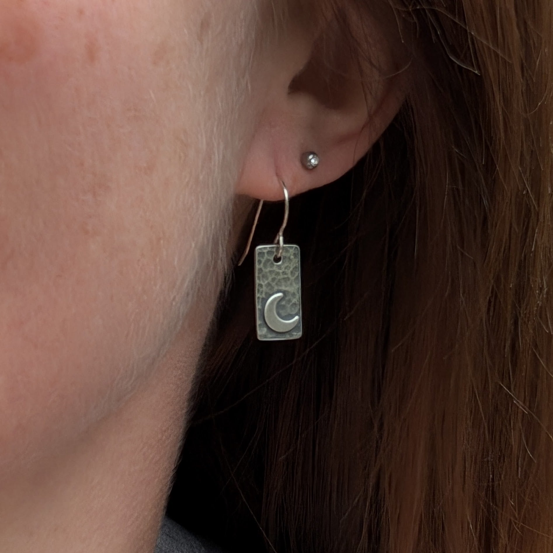 Close-up of an oxidised rectangular earring with a hammered texture and crescent moon detail, shown worn on an ear, highlighting the earring’s size and shape.