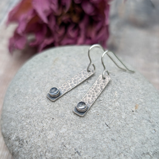 Pair of oxidised sterling silver rectangle earrings with a hammered texture, placed on a smooth stone, each earring featuring a moonstone at the base.