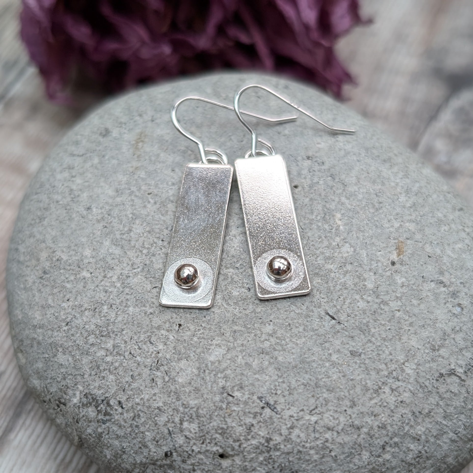 A pair of rectangular silver earrings with a small round silver pebble at the bottom of each rectangle, placed on a smooth gray stone.