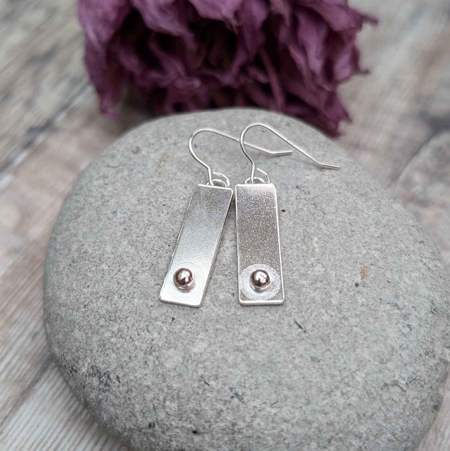 A pair of rectangular silver earrings with a small round silver pebble at the bottom of each rectangle, placed on a smooth gray stone.