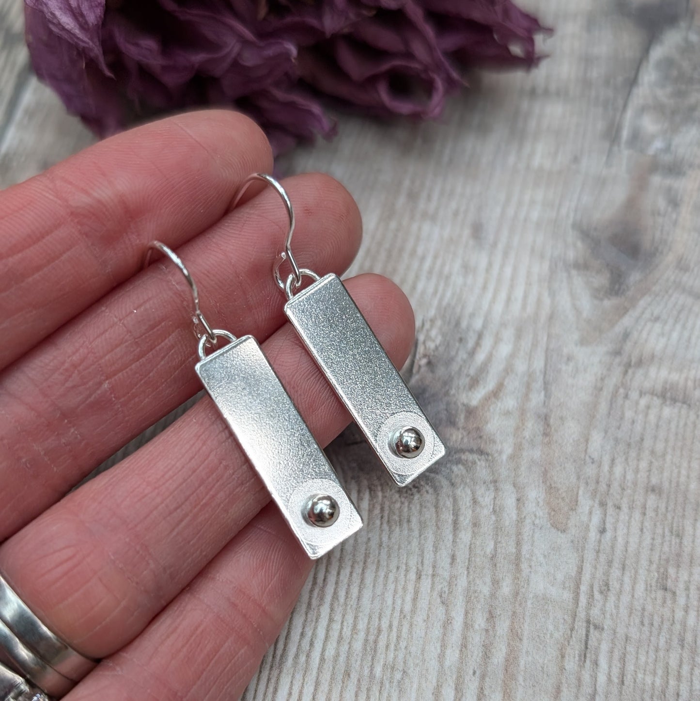 A pair of rectangular silver earrings with a small round silver stud at the bottom of each rectangle, held in a person's hand for size reference, with a wooden background.