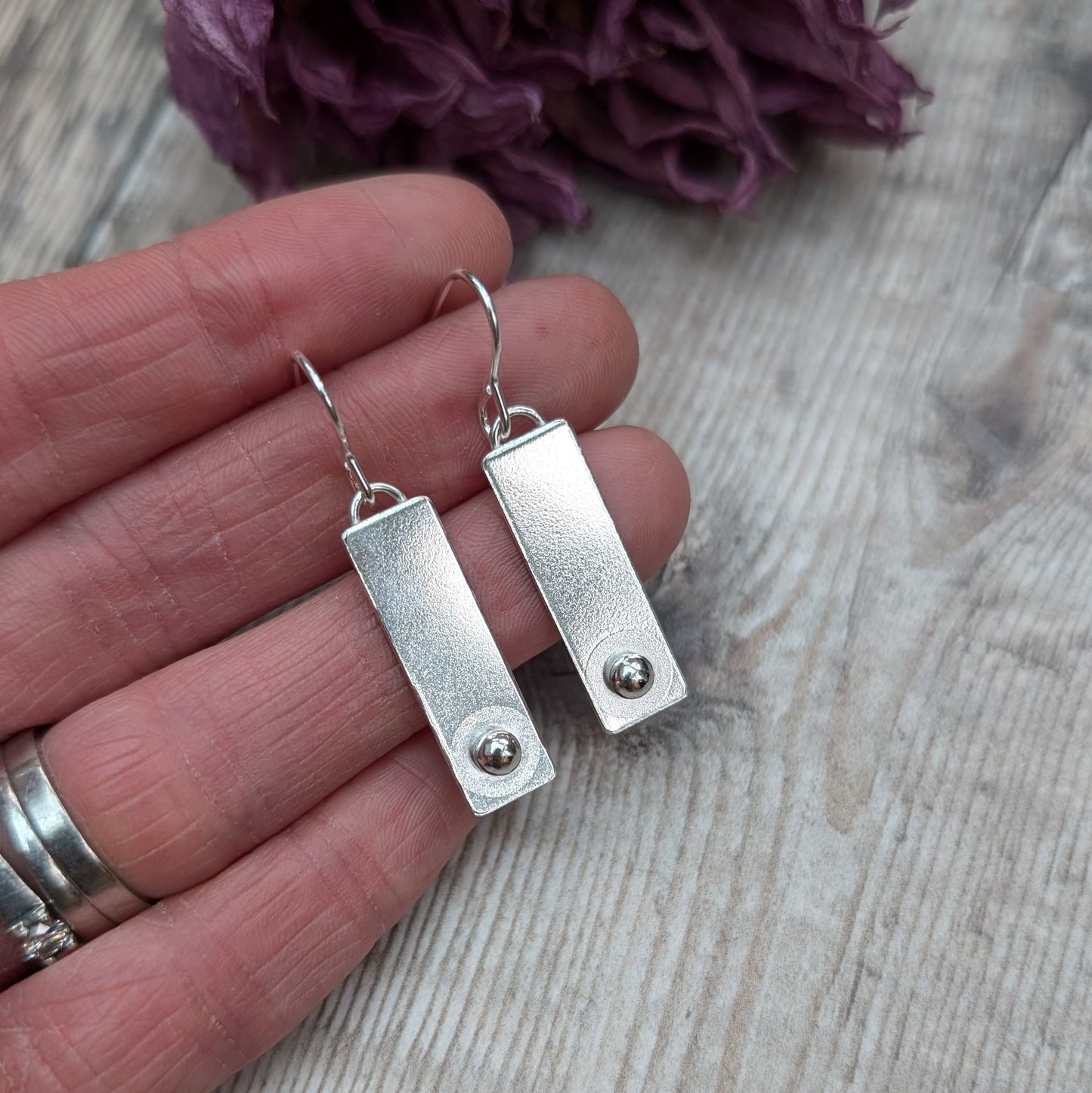 A pair of rectangular silver earrings with a small round silver stud at the bottom of each rectangle, held in a person's hand for size reference, with a wooden background.