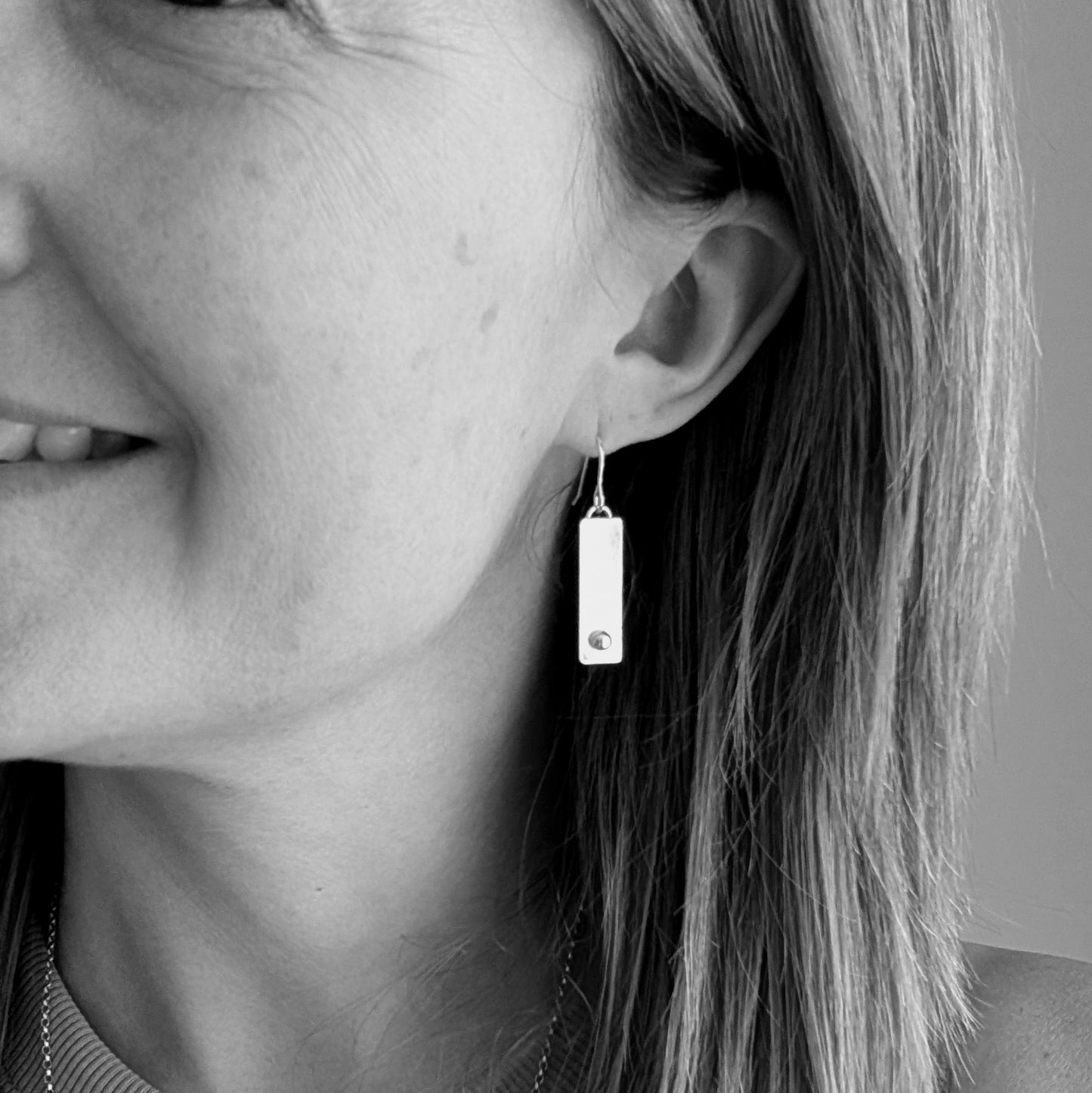 A pair of rectangular silver earrings with a small round silver pebble at the bottom of each rectangle, shown being worn for size reference