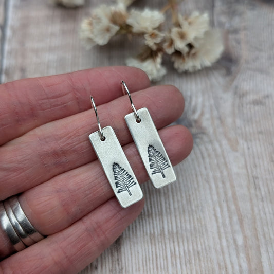 A hand holding a pair of oxidised silver rectangle earrings with an etched evergreen tree design. The earrings' subtle texture and natural theme stand out against the hand's skin, with dried flowers softly blurred in the background.