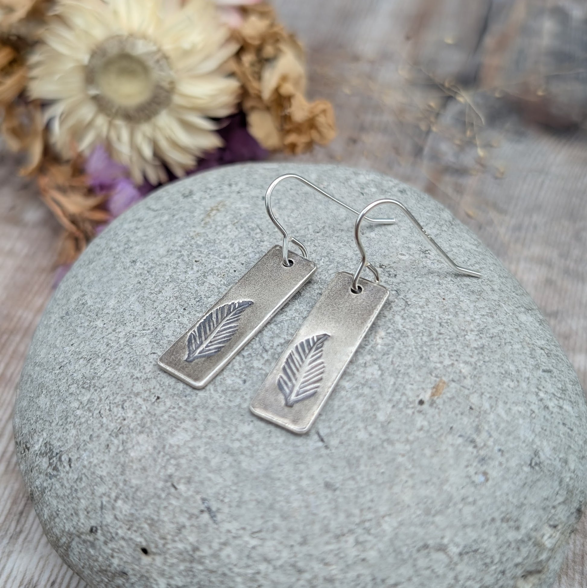 A pair of handmade recycled sterling silver rectangle drop earrings with a hand-stamped feather design, displayed on a smooth grey stone with dried flowers in the background.