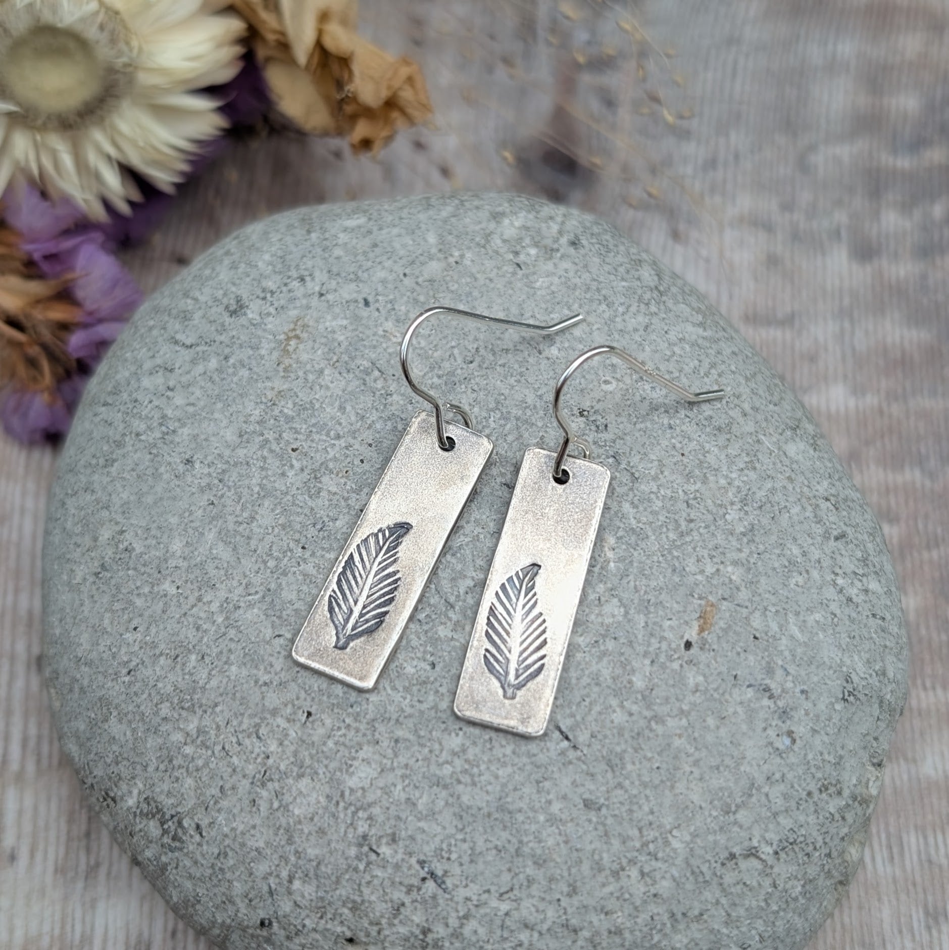 A pair of handmade recycled sterling silver rectangle drop earrings with a hand-stamped feather design, displayed on a smooth grey stone with dried flowers in the background.