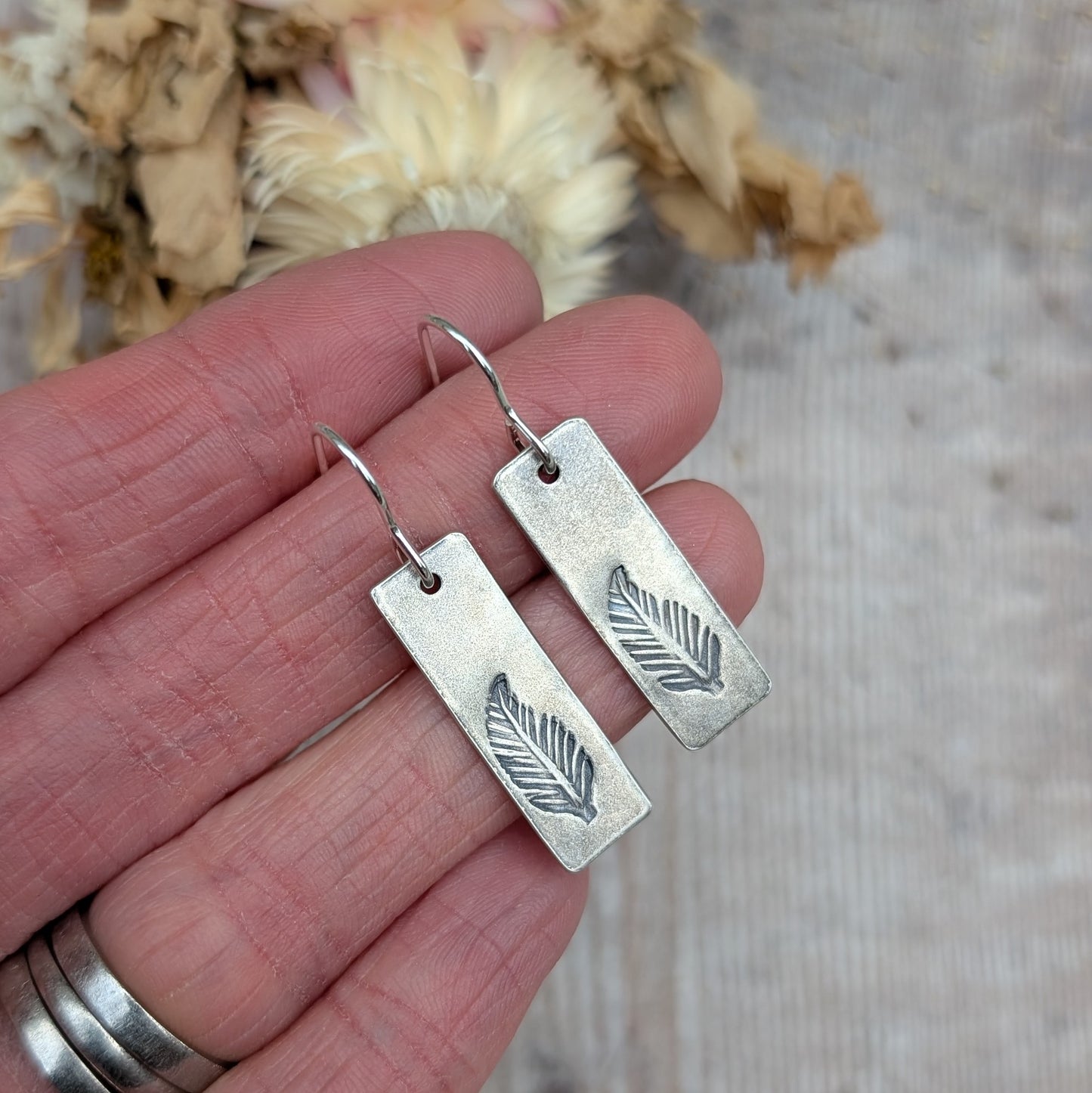 Close-up of a pair of sterling silver rectangle feather earrings being held in a hand, showing their oxidised finish and intricate stamped feather pattern.