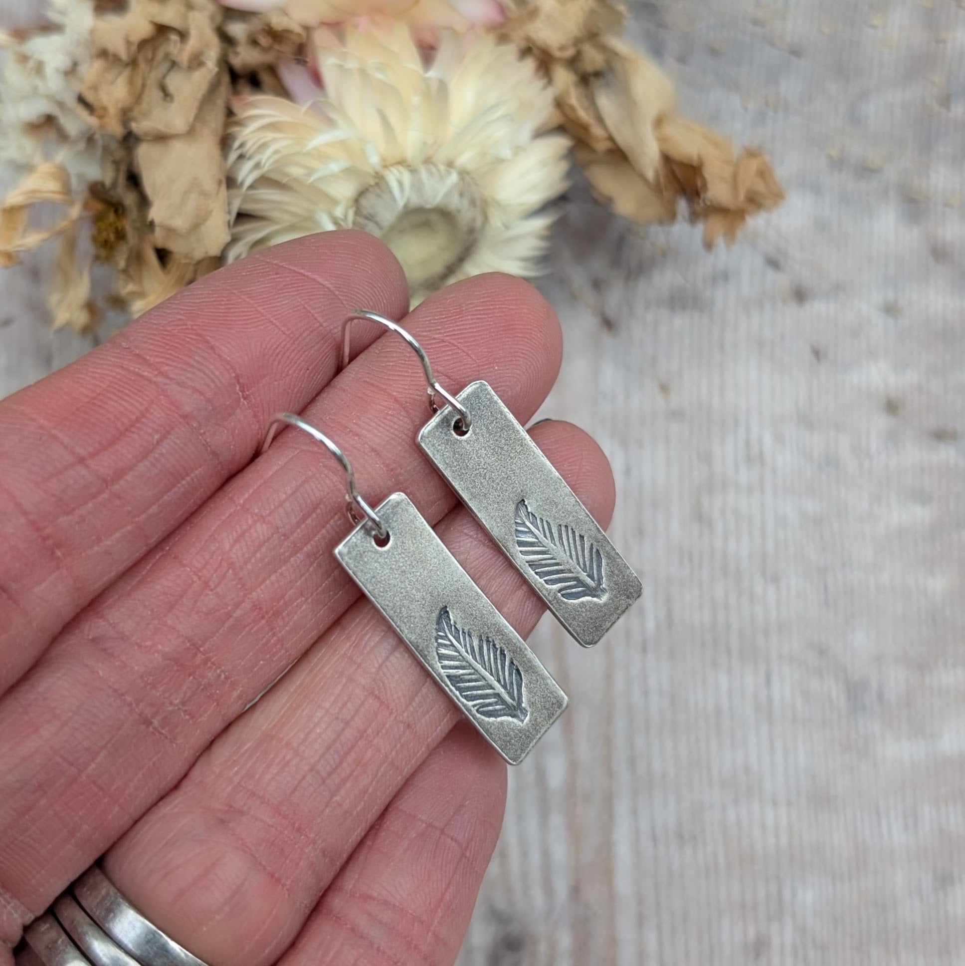 Close-up of a pair of sterling silver rectangle feather earrings being held in a hand, showing their oxidised finish and intricate stamped feather pattern.