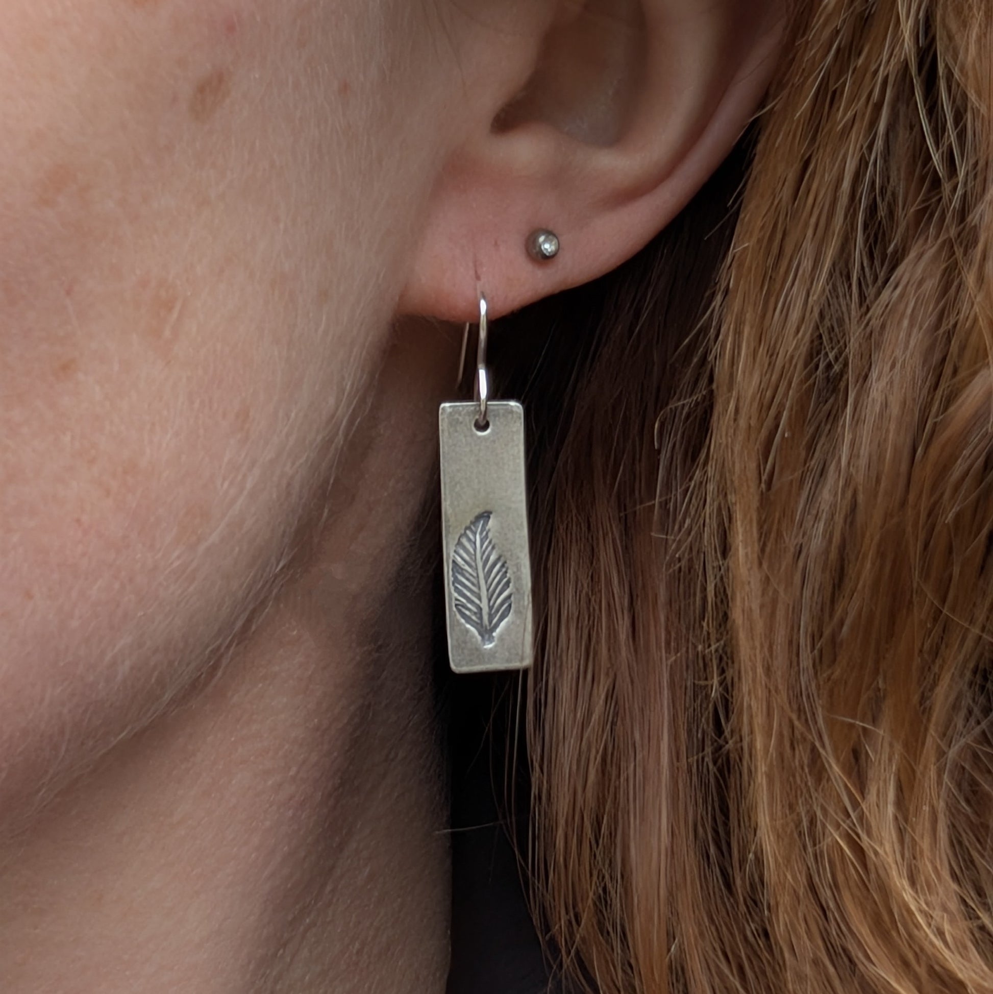 A single rectangular silver earring with a stamped feather design, worn in the ear, showing its length and lightweight style against the wearer's hair.