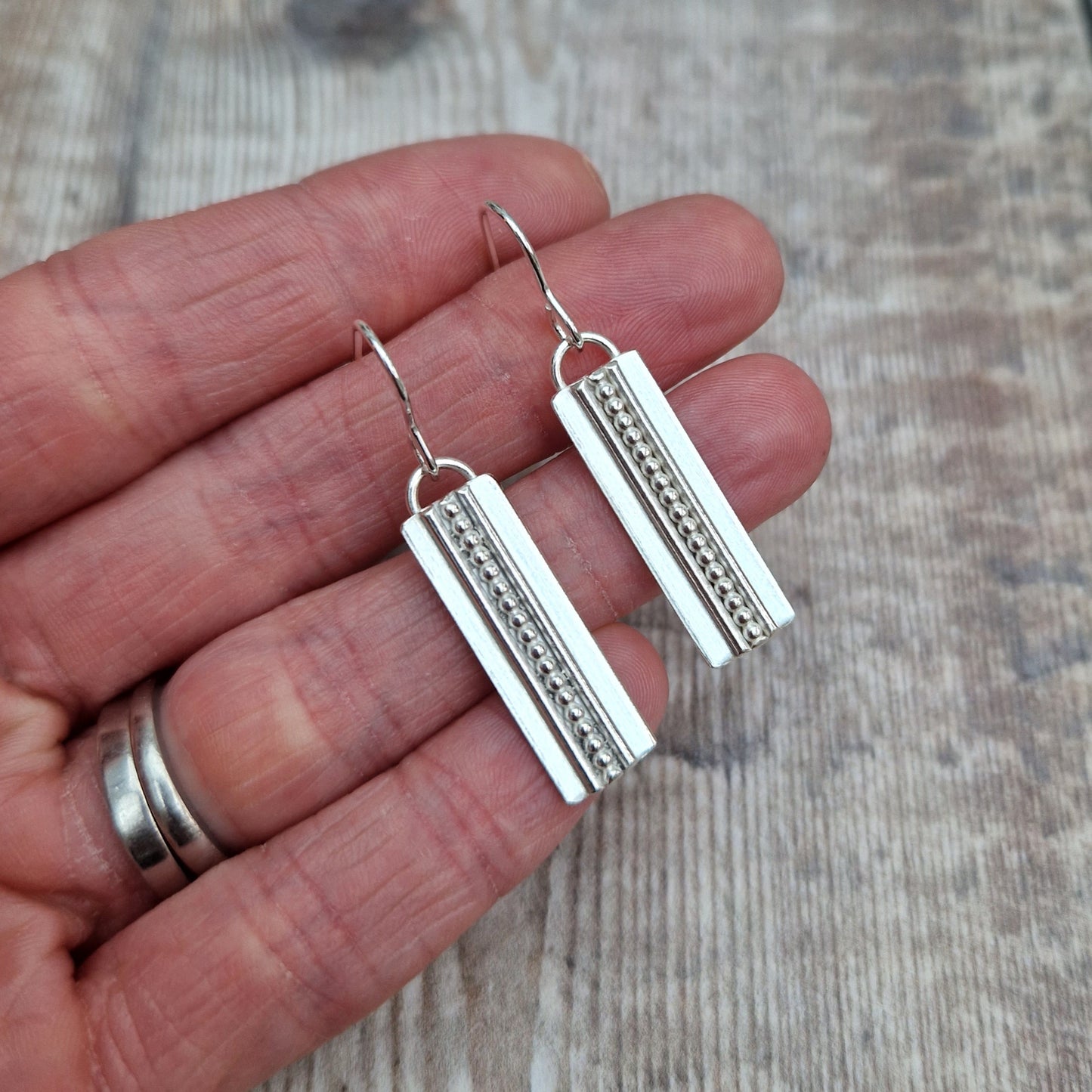 Each earring is a Sterling Silver rectangle suspended from silver earring wire. Along the centre of the rectangle is a raised beaded line with a raised smooth line on each side, running top to bottom of the rectangle.