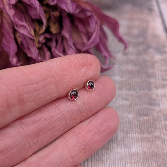 A close-up of a pair of small rhodolite garnet stud earrings set in recycled sterling silver, resting on a person's fingers. The deep red gemstones catch the light against a backdrop of dried purple flowers and a rustic wooden surface.