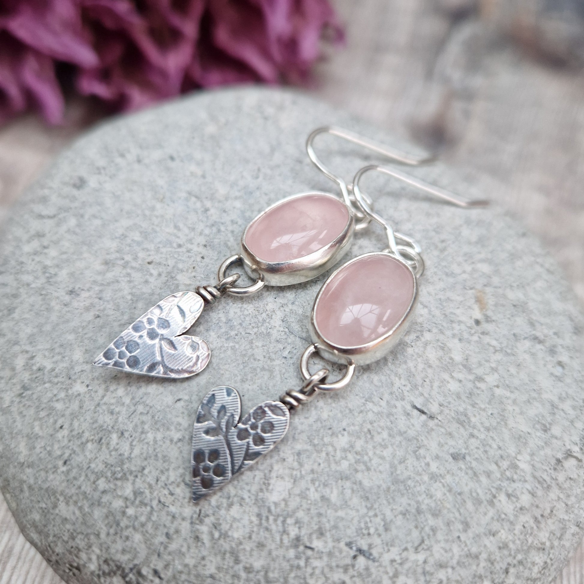 Each earring is an oval pink Rose Quartz Gemstone set in a Sterling Silver bezel suspended from earring wire via a silver D link. Suspended from the Rose Quartz Gemstone is a silver heart shape with stamped floral design.