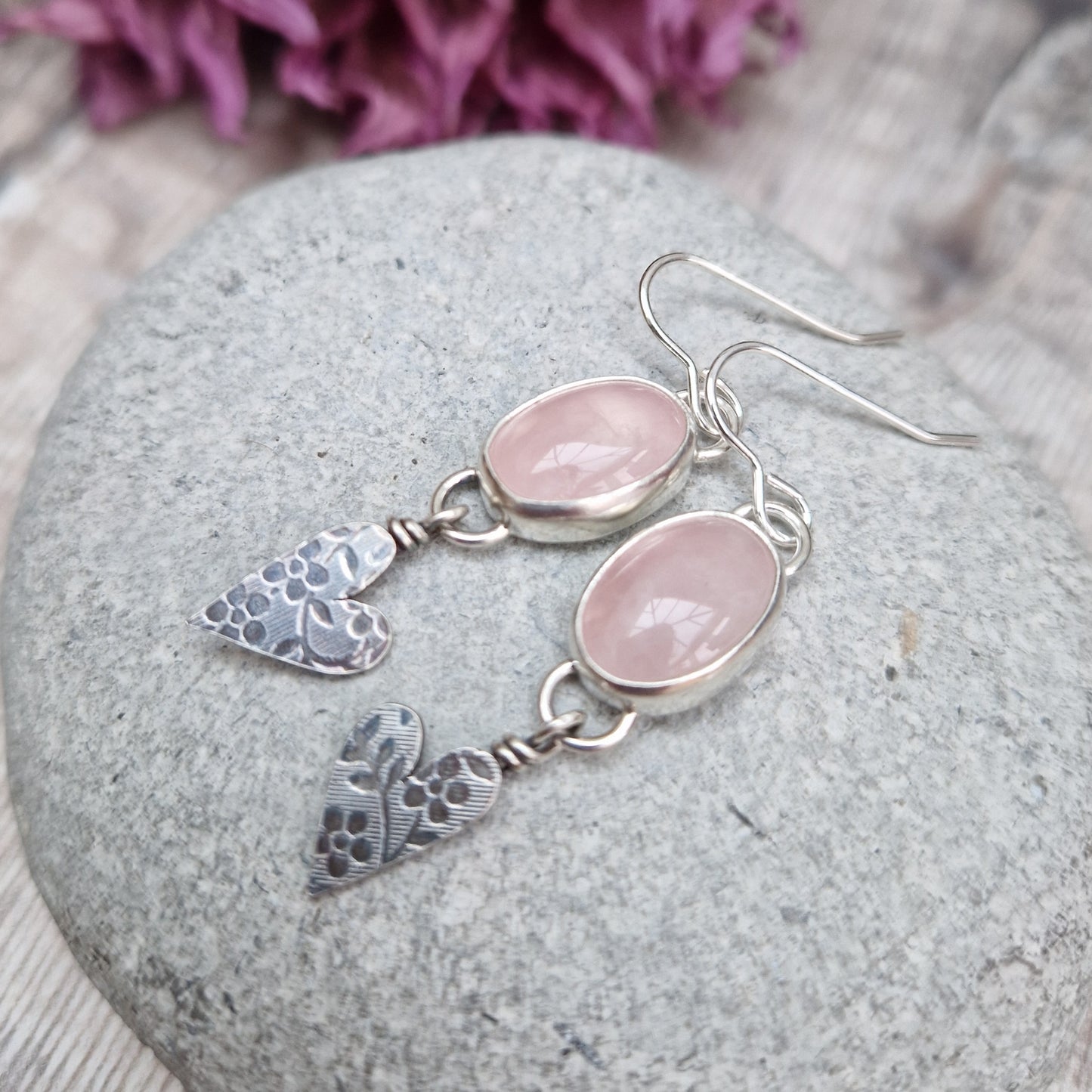 Each earring is an oval pink Rose Quartz Gemstone set in a Sterling Silver bezel suspended from earring wire via a silver D link. Suspended from the Rose Quartz Gemstone is a silver heart shape with stamped floral design.