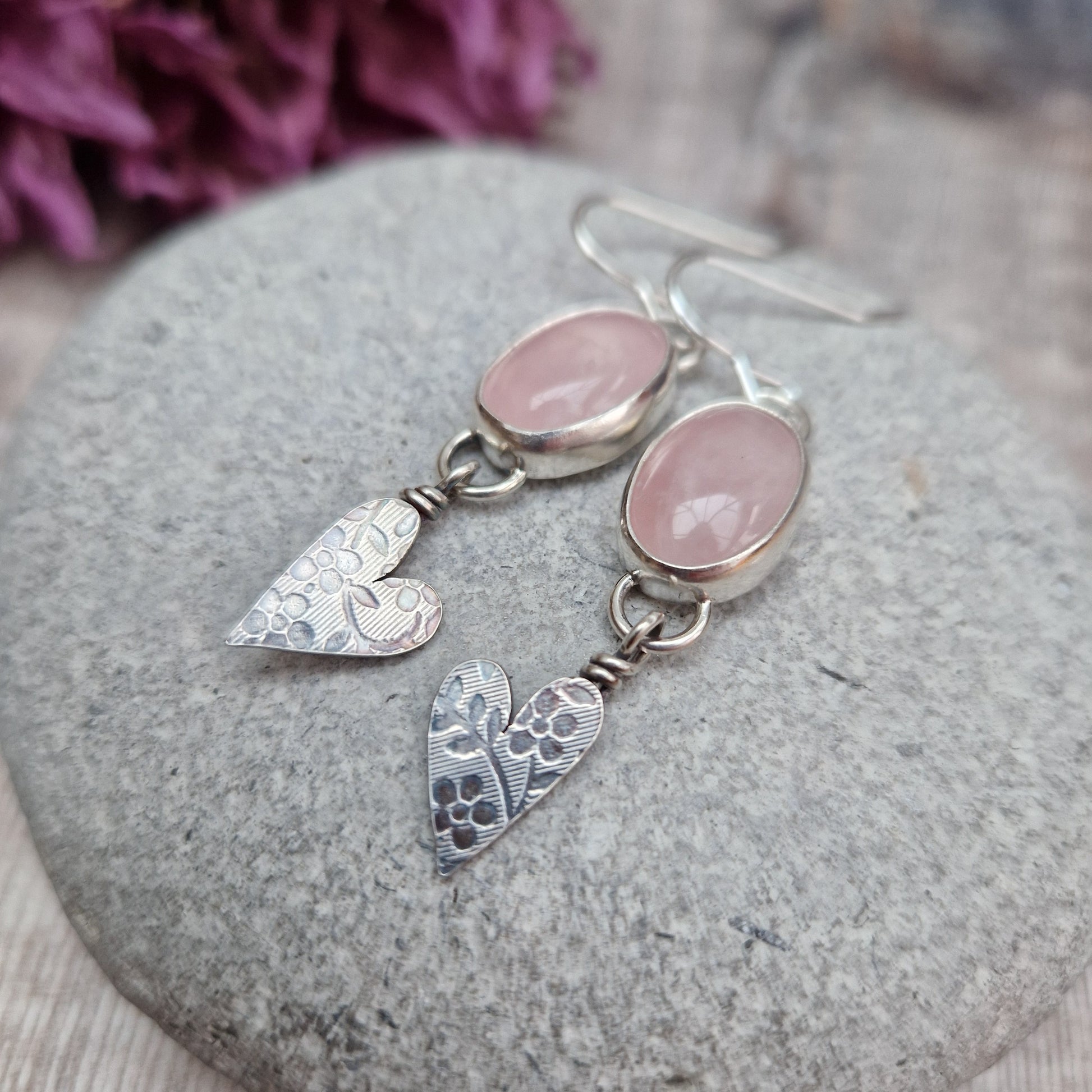 Each earring is an oval pink Rose Quartz Gemstone set in a Sterling Silver bezel suspended from earring wire via a silver D link. Suspended from the Rose Quartz Gemstone is a silver heart shape with stamped floral design.