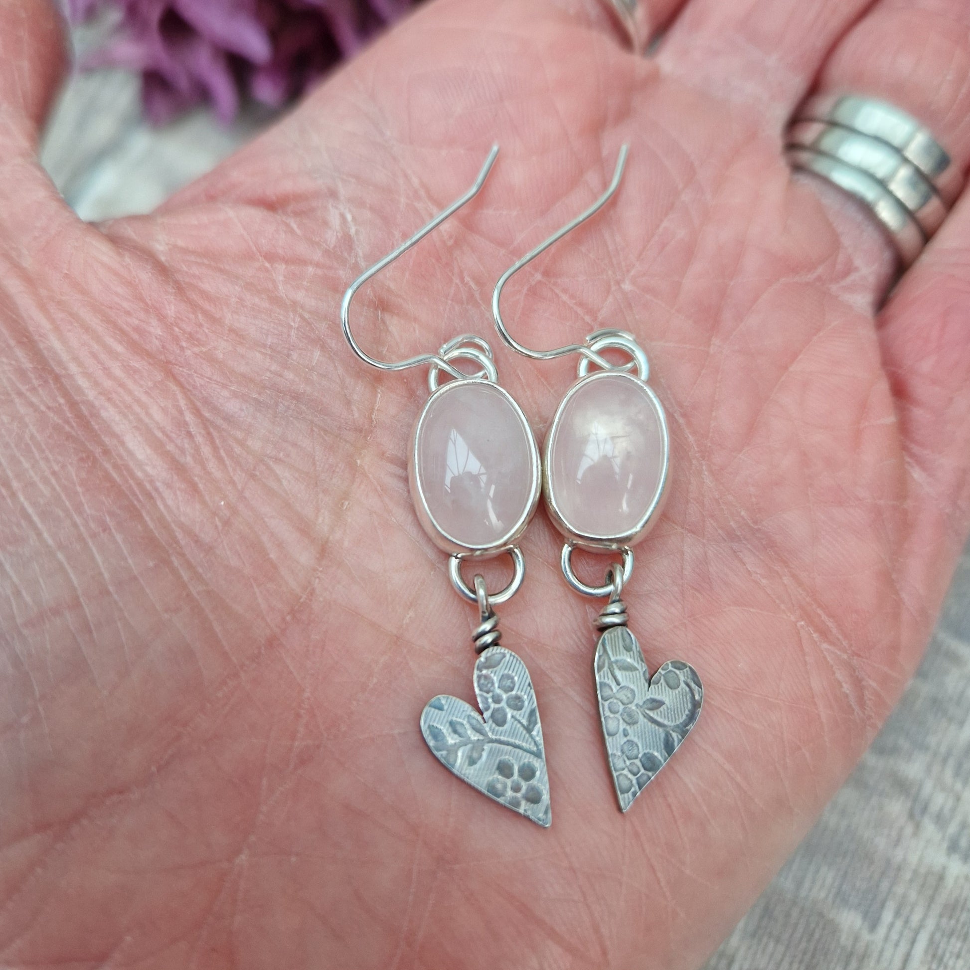 Each earring is an oval pink Rose Quartz Gemstone set in a Sterling Silver bezel suspended from earring wire via a silver D link. Suspended from the Rose Quartz Gemstone is a silver heart shape with stamped floral design.