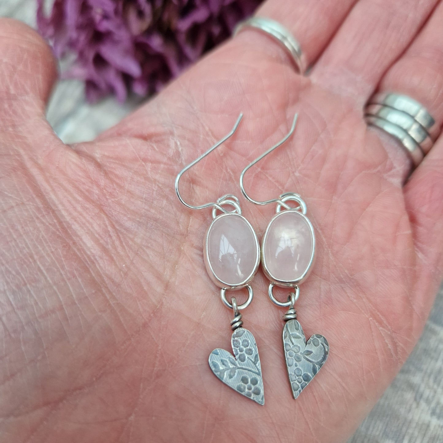 Each earring is an oval pink Rose Quartz Gemstone set in a Sterling Silver bezel suspended from earring wire via a silver D link. Suspended from the Rose Quartz Gemstone is a silver heart shape with stamped floral design.