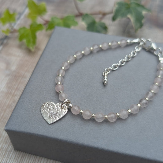 Handmade rose quartz and silver floral heart charm bracelet photographed on a grey jewellery box with ivy in the background, showing the textured sterling silver heart charm and pale pink rose quartz gemstone beads.
