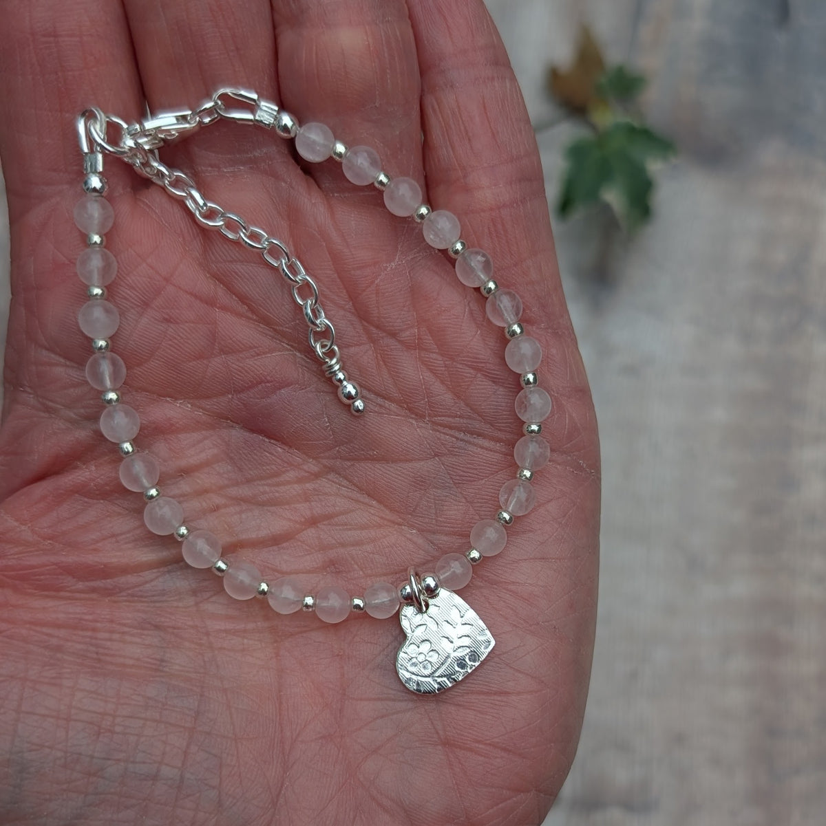 Rose quartz and silver floral heart charm bracelet displayed in the palm of a hand, showing pale pink rose quartz beads, sterling silver spacers, a floral heart charm and an adjustable silver clasp.