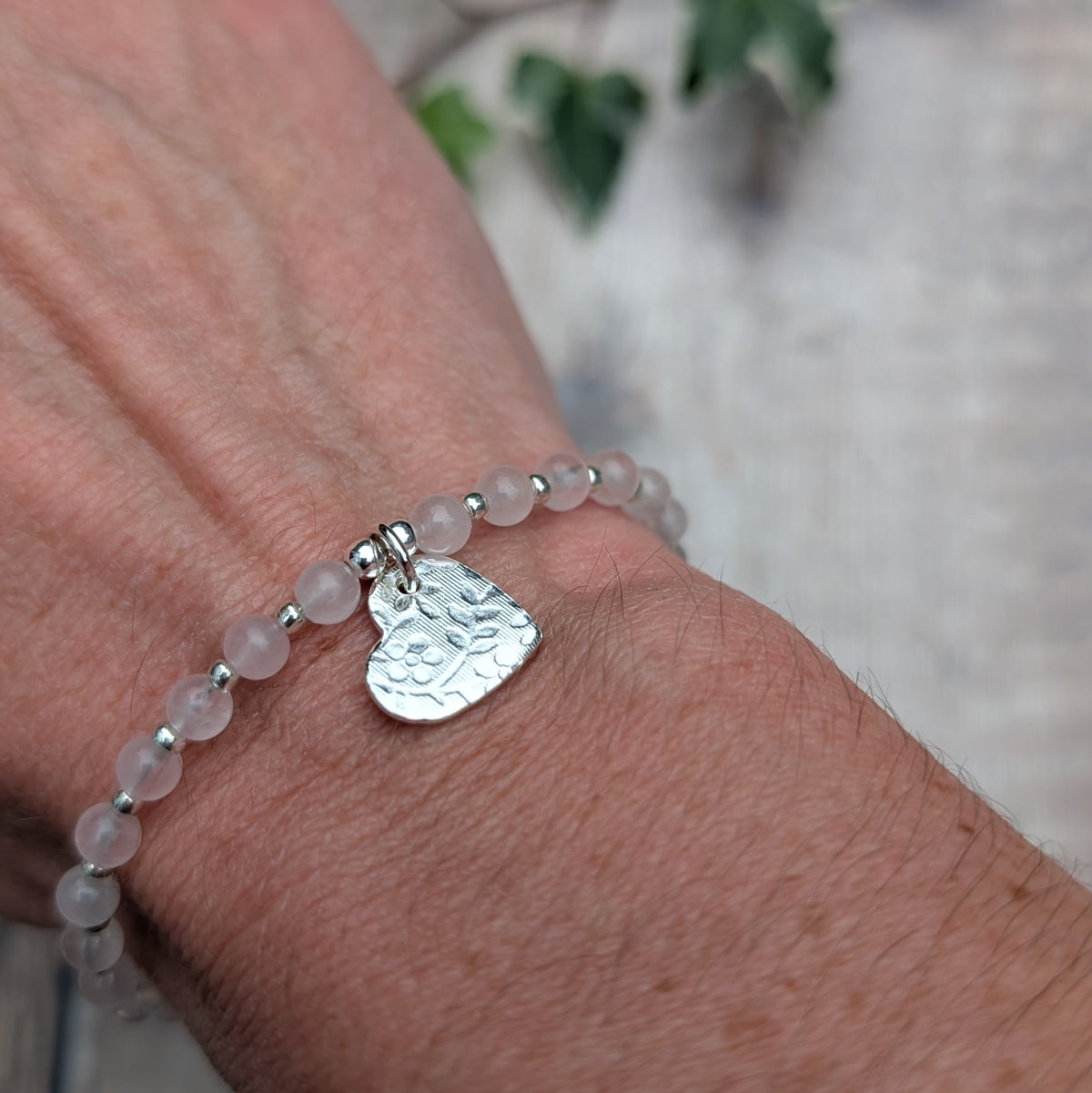 Rose quartz and silver floral heart charm bracelet being worn on a wrist, showing the textured silver heart charm hanging from the soft pink rose quartz beads.