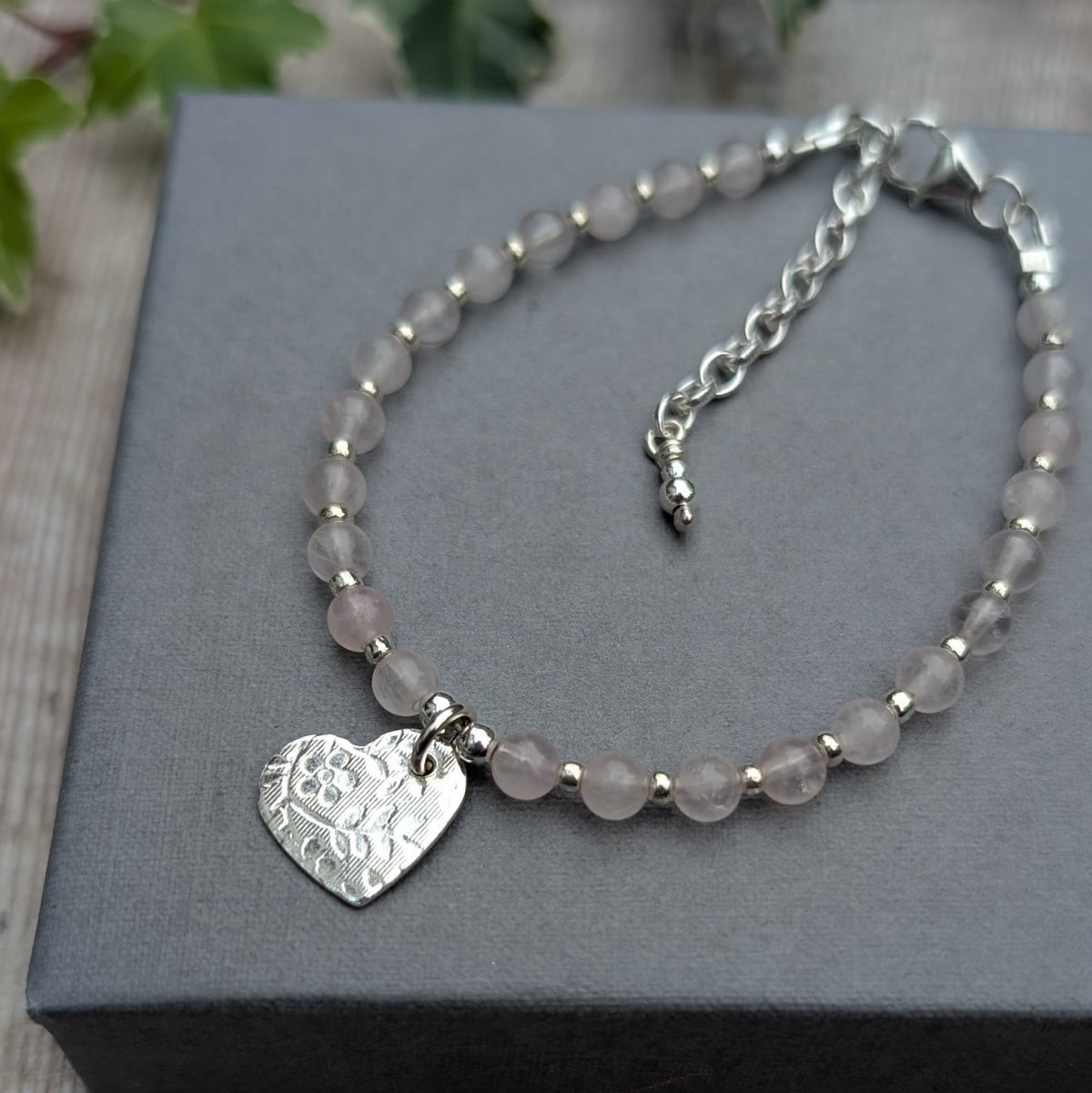 Rose quartz and silver floral heart charm bracelet displayed on a grey jewellery box, featuring pale pink rose quartz beads, small silver spacers, a floral patterned heart charm and an adjustable extension chain.