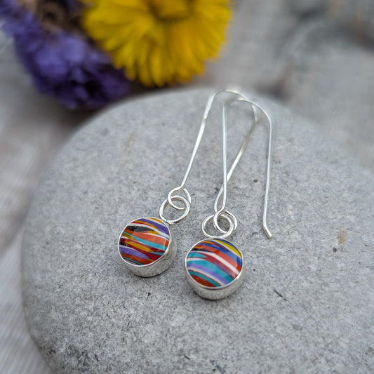 A pair of long silver dangle earrings with round Surfite stones, shown resting on a smooth grey pebble with yellow and purple flowers in the background.
