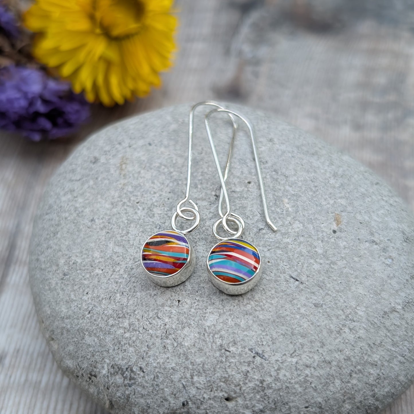 A pair of long silver dangle earrings with round Surfite stones, shown resting on a smooth grey pebble with yellow and purple flowers in the background.