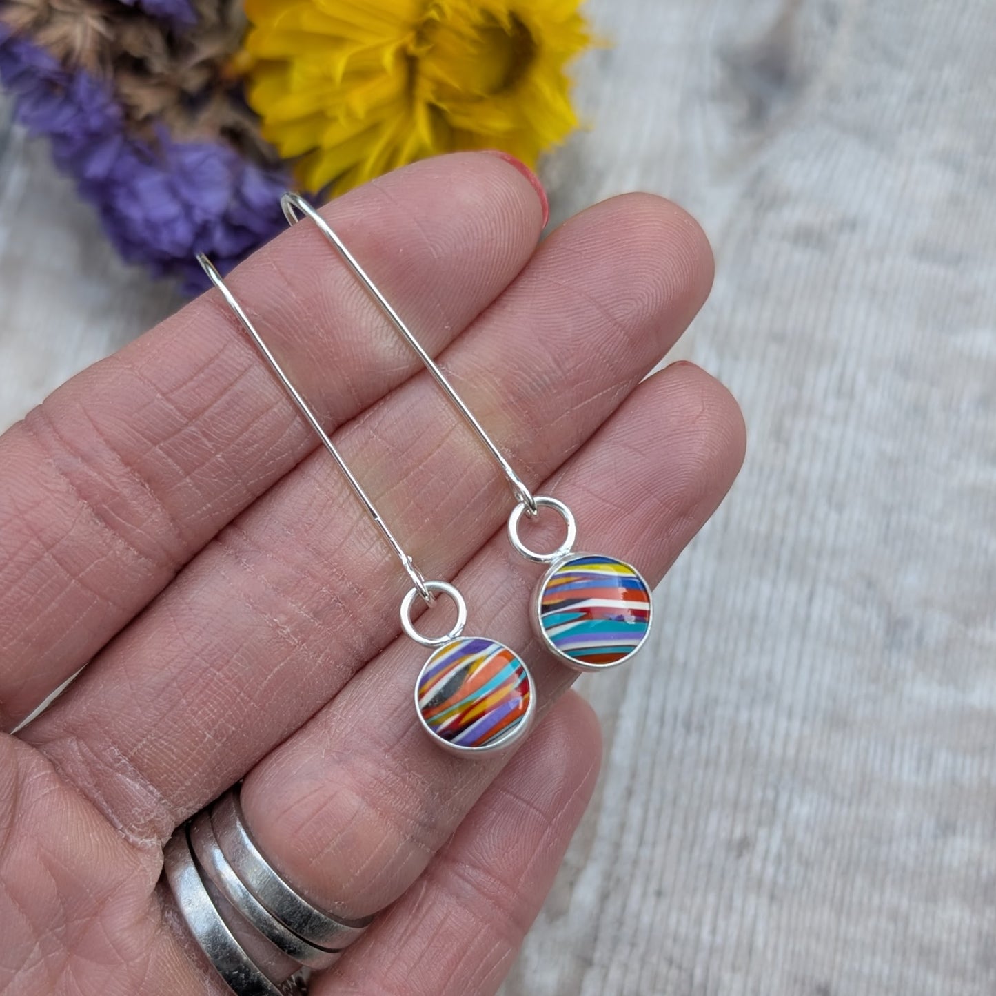 A close-up of a hand holding a pair of long sterling silver earrings featuring round, colourful Surfite stones set in silver bezels.
