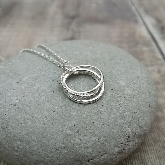 A close-up of a handmade silver necklace featuring three intertwined rings, each with a different texture, lying on a smooth stone surface. One ring is beaded, one is hammered, and the third has a smooth finish.