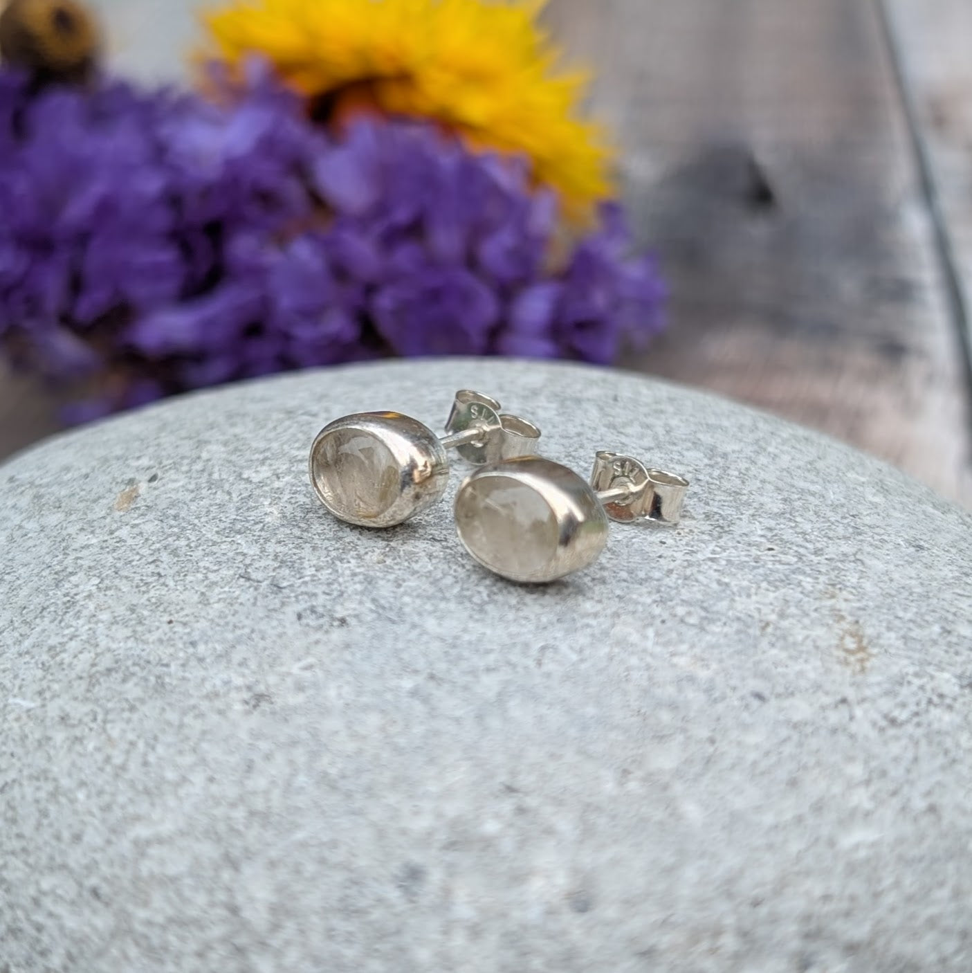 A pair of oval rutile quartz stud earrings resting on a smooth stone, surrounded by blurred purple and yellow flowers in the background. The quartz stones showcase subtle golden inclusions.