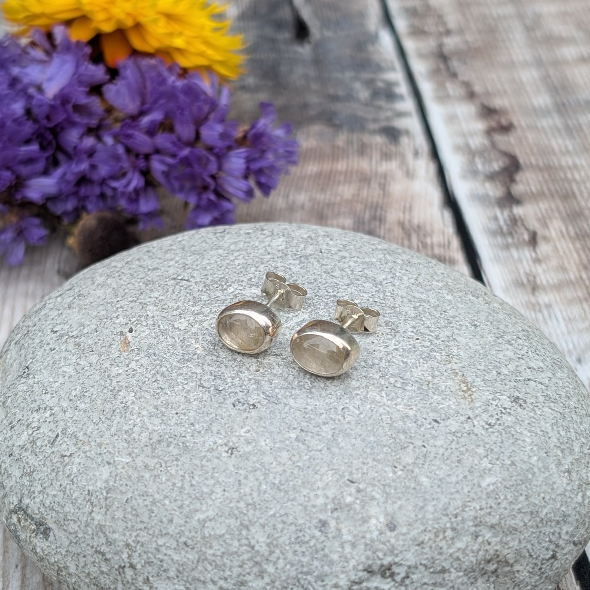 A pair of oval rutile quartz stud earrings resting on a smooth stone, surrounded by blurred purple and yellow flowers in the background. The quartz stones showcase subtle golden inclusions.