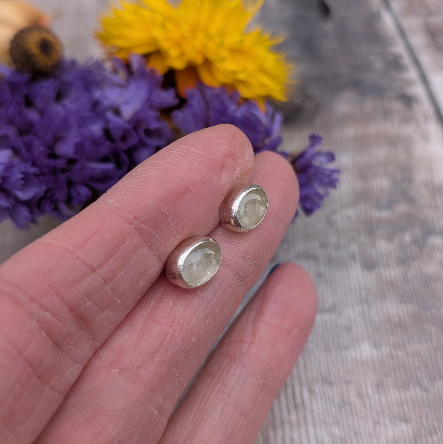 A pair of oval rutile quartz stud earrings set in recycled sterling silver, displayed on a hand with vibrant purple and yellow flowers in the background. The earrings feature golden inclusions within the quartz stones.