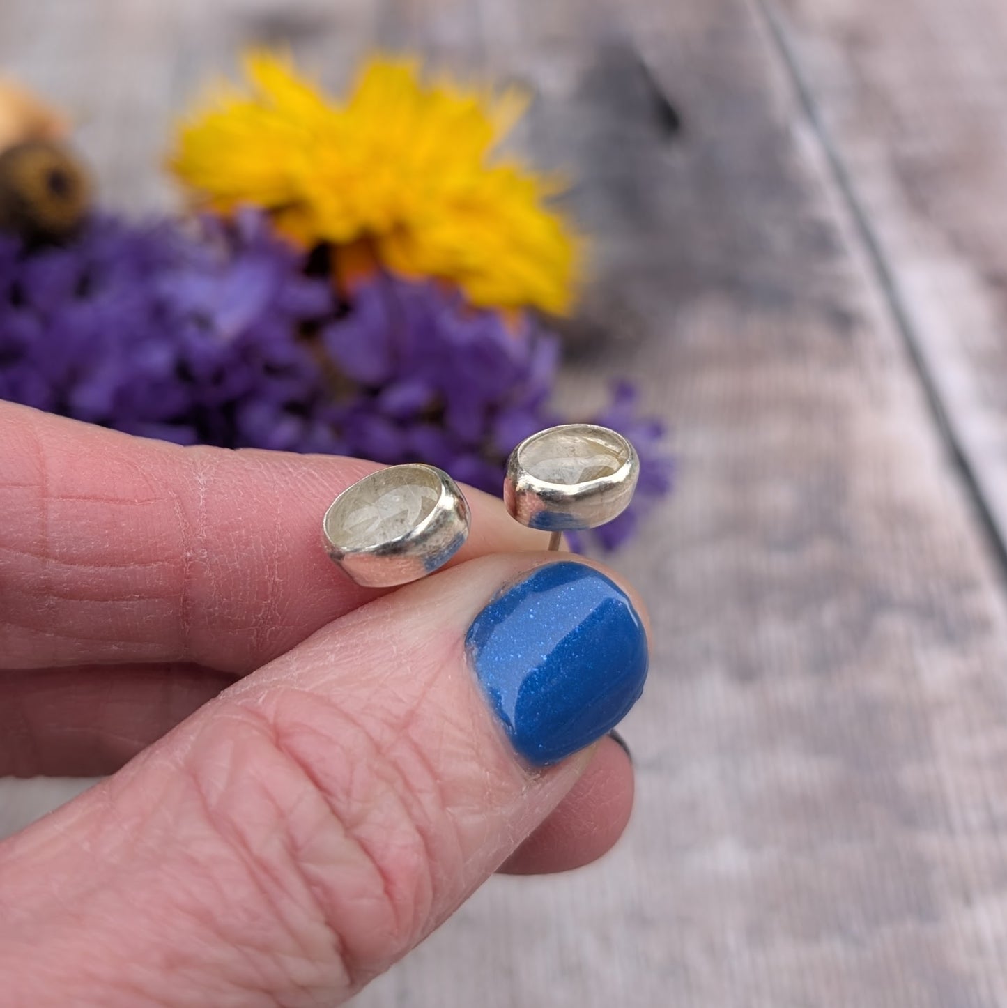 A pair of oval rutile quartz stud earrings set in recycled sterling silver, displayed on a hand with vibrant purple and yellow flowers in the background. The earrings feature golden inclusions within the quartz stones.