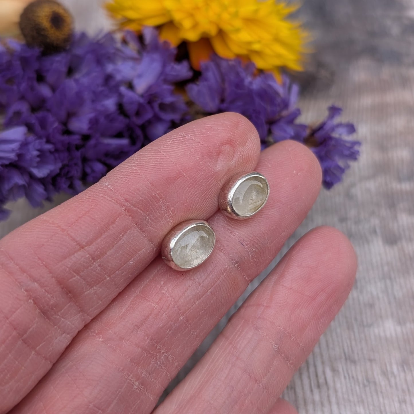 A pair of oval rutile quartz stud earrings set in recycled sterling silver, displayed on a hand with vibrant purple and yellow flowers in the background. The earrings feature golden inclusions within the quartz stones.